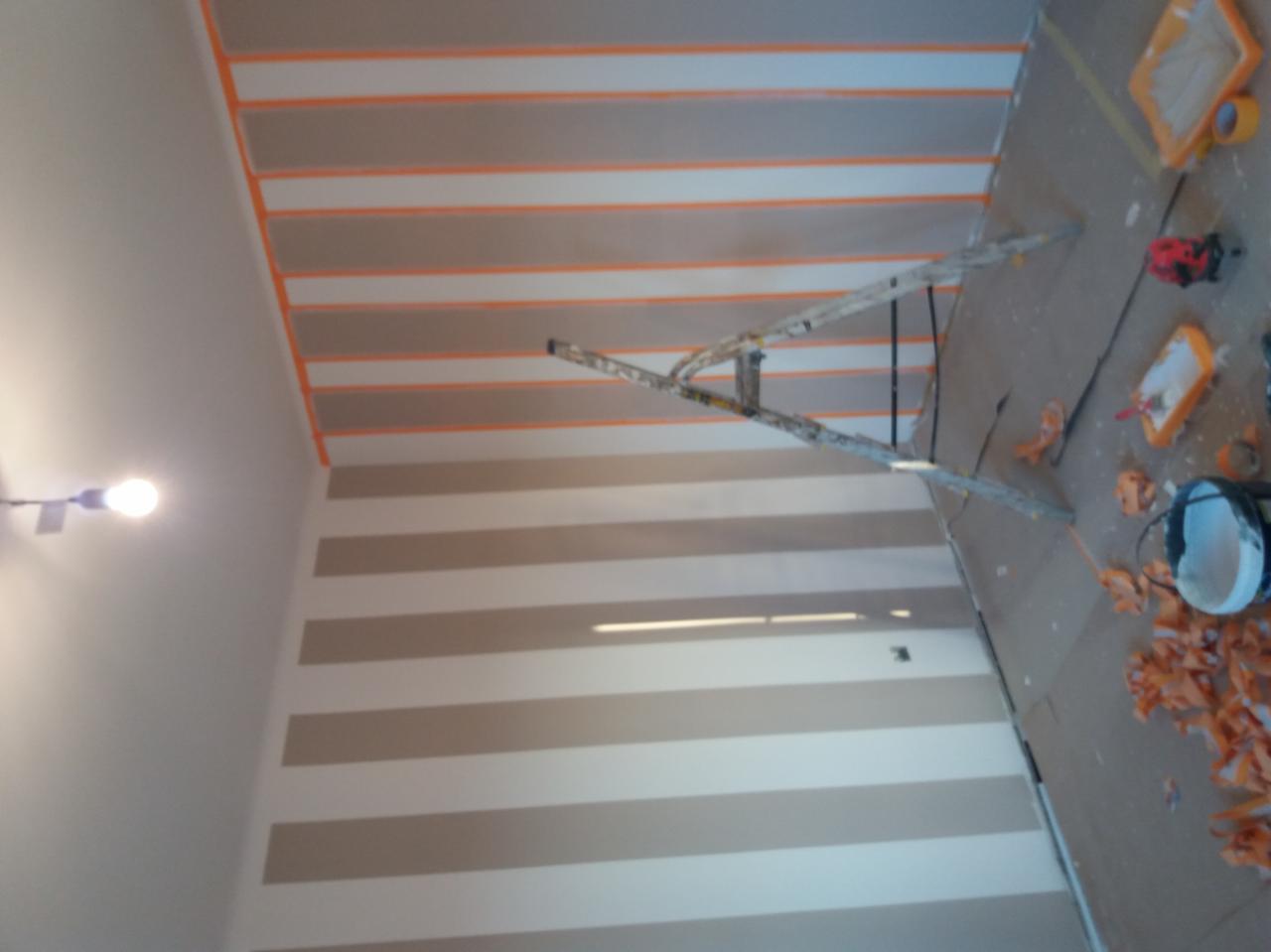 Ściana w trakcie malowania w poziome pasy, zabezpieczona taśmą malarską, z widoczną drabiną, kuwetami malarskimi i resztkami taśmy na podłodze.