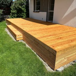 Sebastian Purschel - Nowy, drewniany taras przydomowy z dwustopniowymi schodami, otoczony zielonym trawnikiem i jasną elewacją budynku.