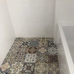 Sebastian Purschel - Wnętrze łazienki z dekoracyjną, wzorzystą podłogą z płytek ceramicznych. Białe, gładkie płytki na ścianach i fragment kabiny prysznicowej.