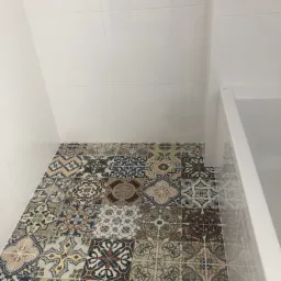 Wnętrze łazienki z dekoracyjną, wzorzystą podłogą z płytek ceramicznych. Białe, gładkie płytki na ścianach i fragment kabiny prysznicowej.