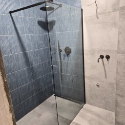 Sebastian Purschel - Kabina prysznicowa walk-in z czarną ramą i deszczownicą, wykończona niebieską mozaiką i szarą płytką. Widoczne baterie w kolorze czarnym. Minimalistyczny design.