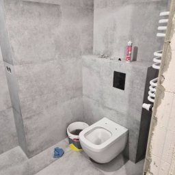 Sebastian Purschel - Szara łazienka w trakcie wykańczania: podwieszana toaleta, grzejnik drabinkowy, płytki imitujące beton, wiadro i akcesoria montażowe. Widoczne ślady prac wykończeniowych.