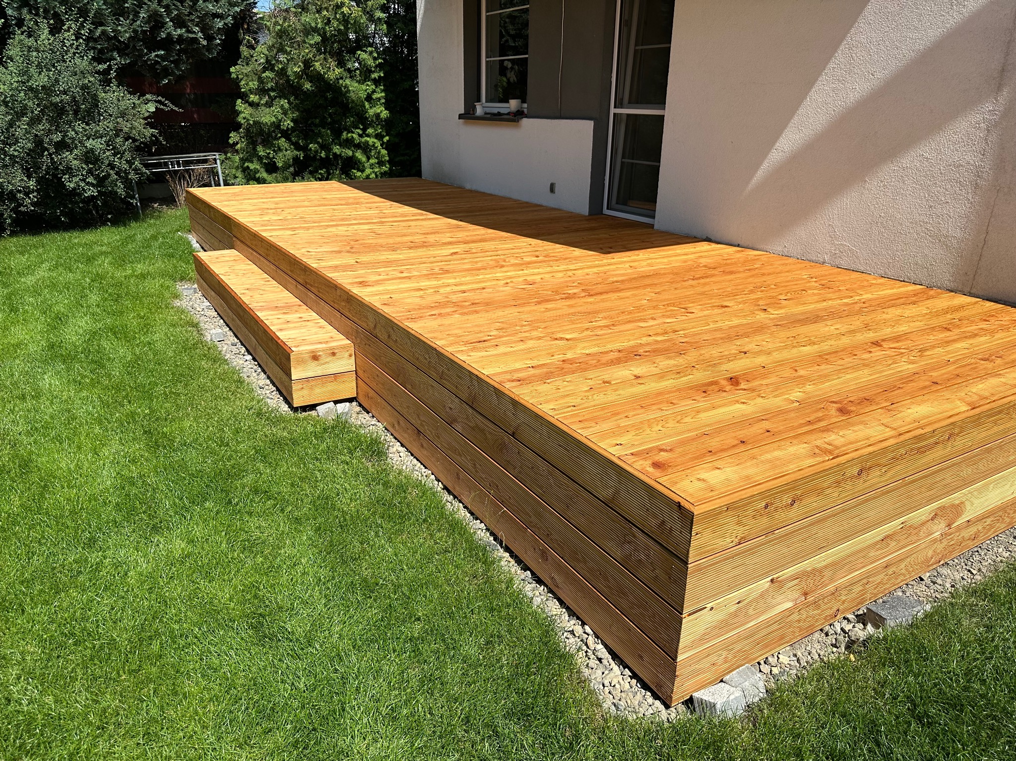 Nowy, drewniany taras przydomowy z dwustopniowymi schodami, otoczony zielonym trawnikiem i jasną elewacją budynku.