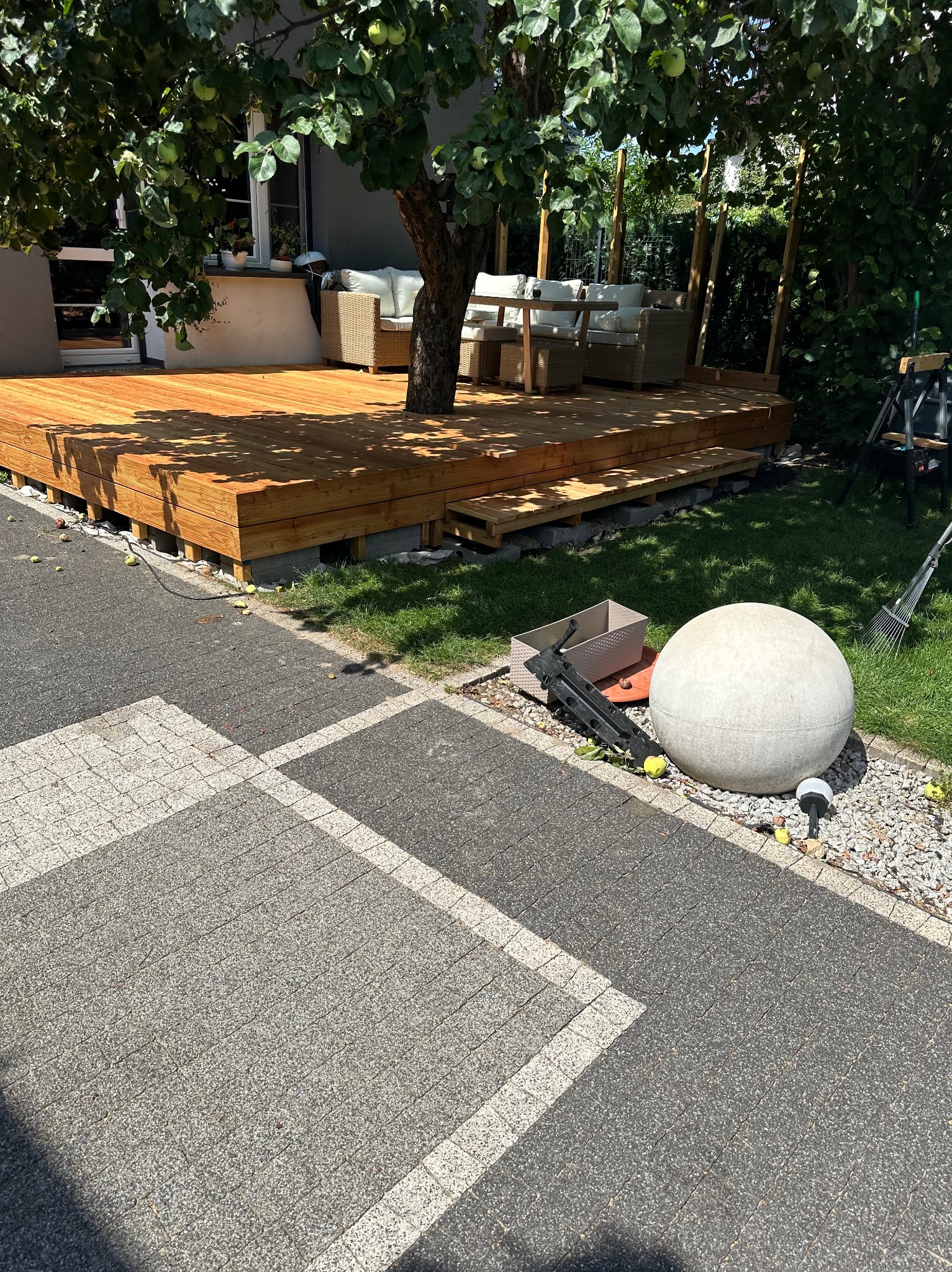 Nowy drewniany taras z meblami ogrodowymi pod drzewem, widok z poziomu kostki brukowej. Na pierwszym planie dekoracyjna kula ogrodowa i narzędzia.