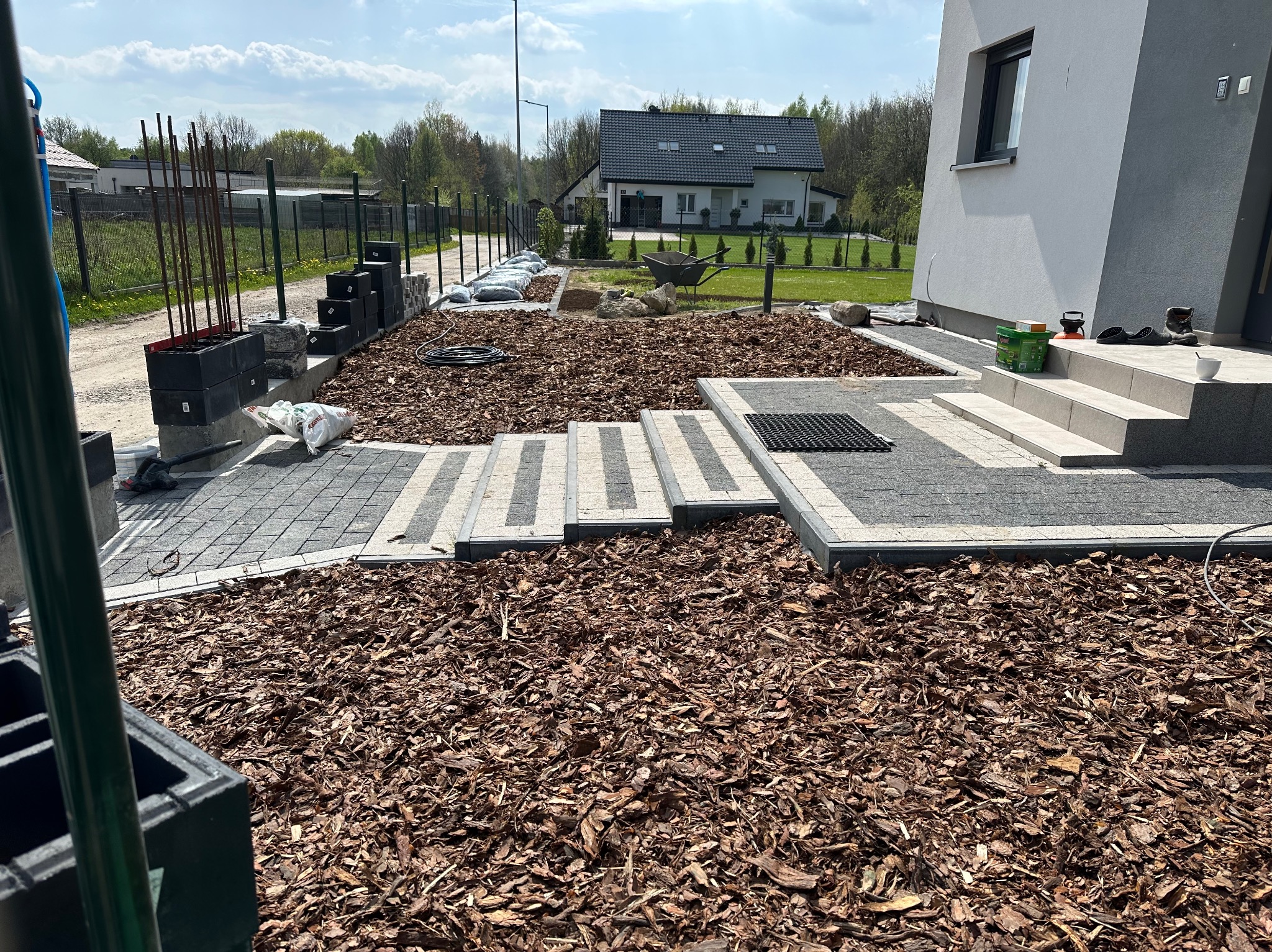 Układanie kostki brukowej i kory na podwórku z widokiem na dom w słoneczny dzień, widoczne materiały budowlane i narzędzia.