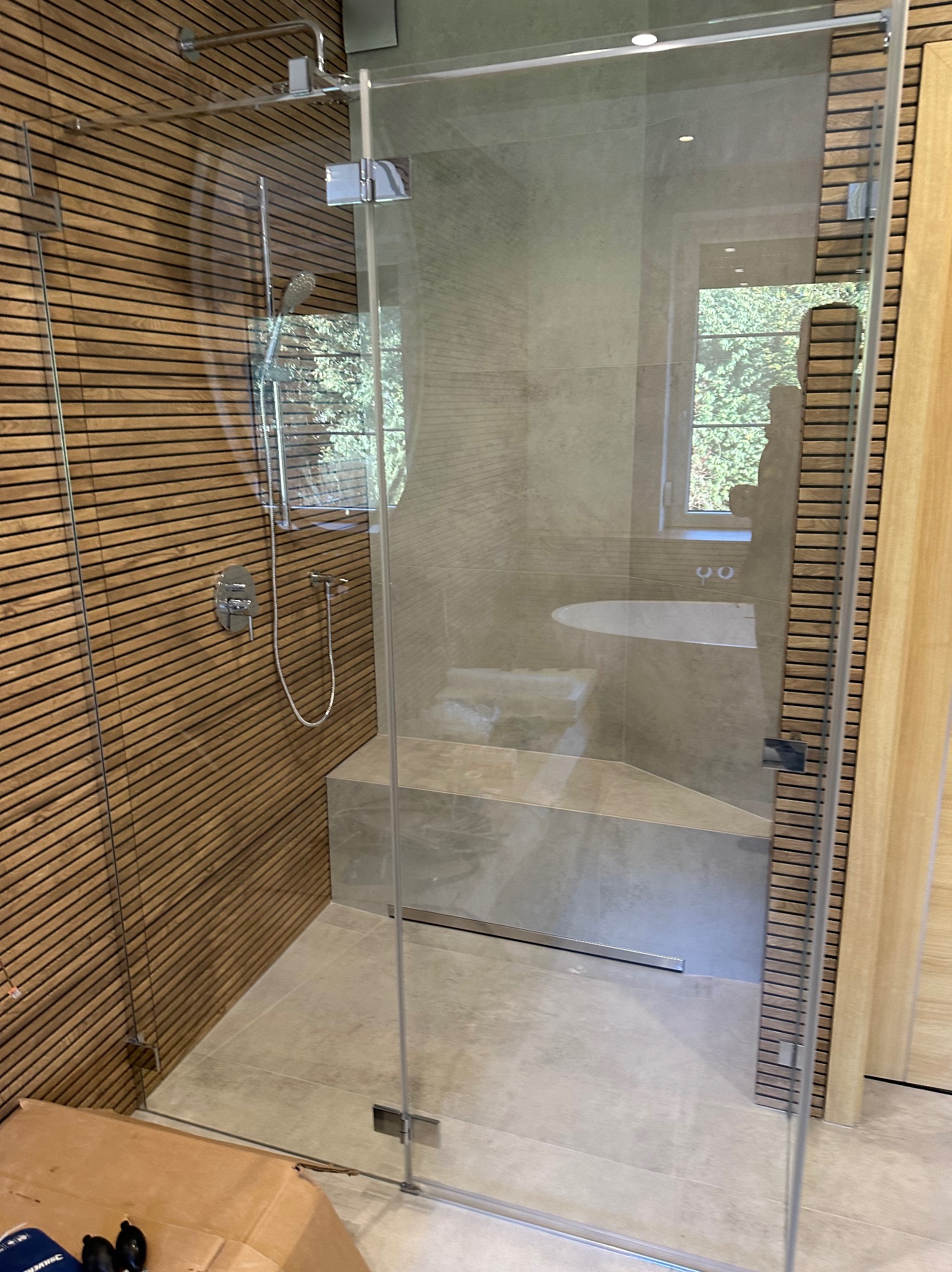 Nowoczesna kabina prysznicowa z drewnianą ścianą, szklanymi drzwiami i siedziskiem, widoczna bateria i słuchawka prysznicowa, w tle wanna i okno