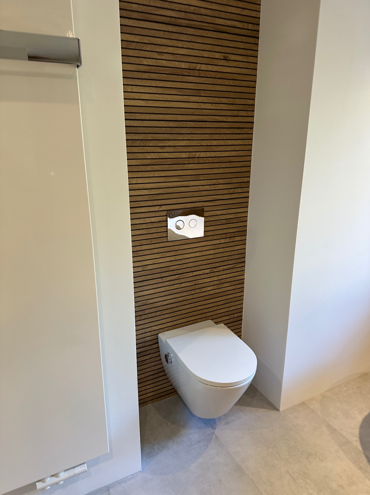 Nowoczesna toaleta podwieszana z chromowanym przyciskiem spłukującym na tle ściany z drewnianych paneli, jasna podłoga z płytek.