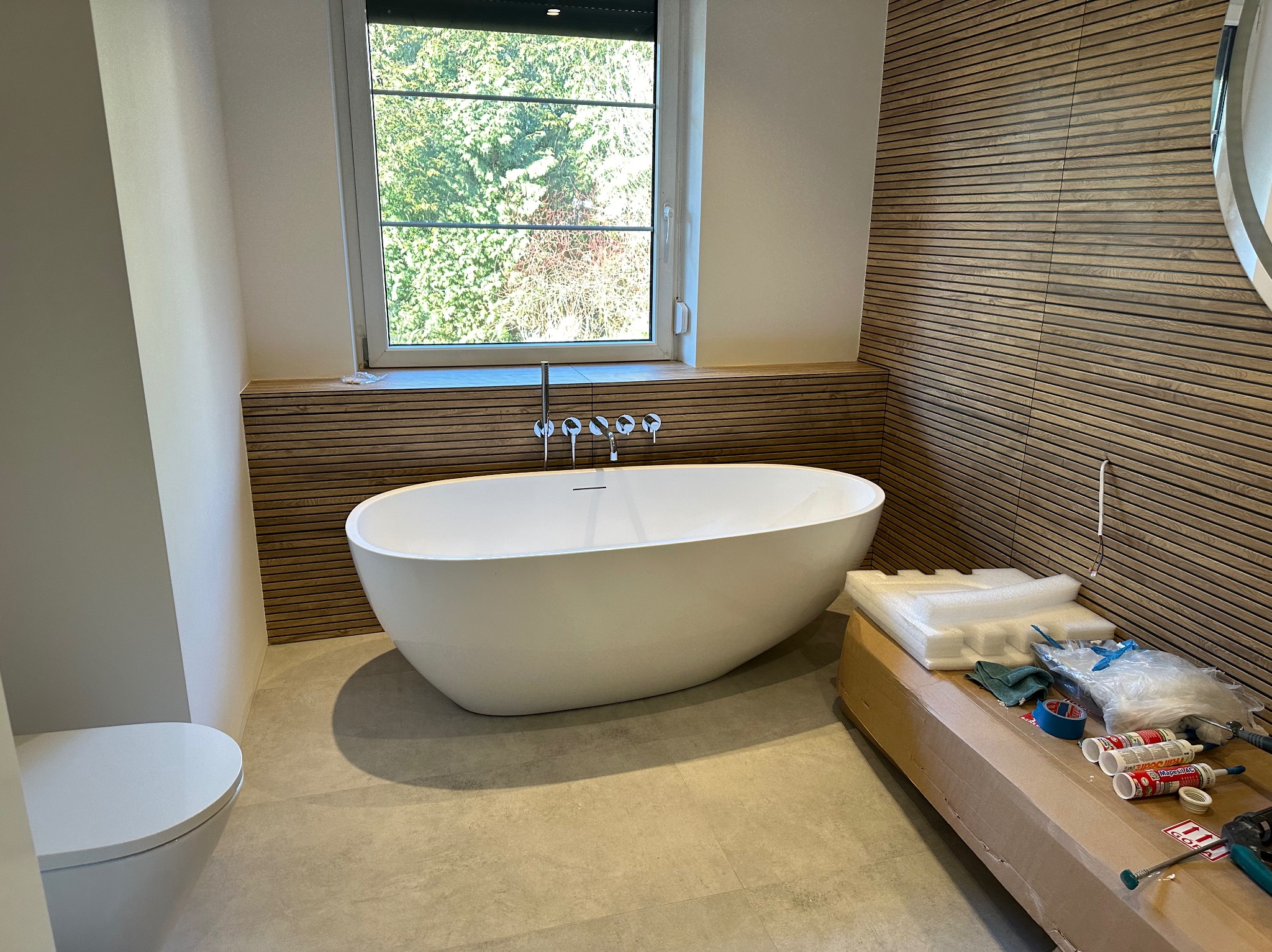 Nowoczesna łazienka w trakcie wykończenia: wolnostojąca wanna, drewniana okładzina ścienna, okno z widokiem na zieleń, karton z materiałami budowlanymi i narzędziami w tle.