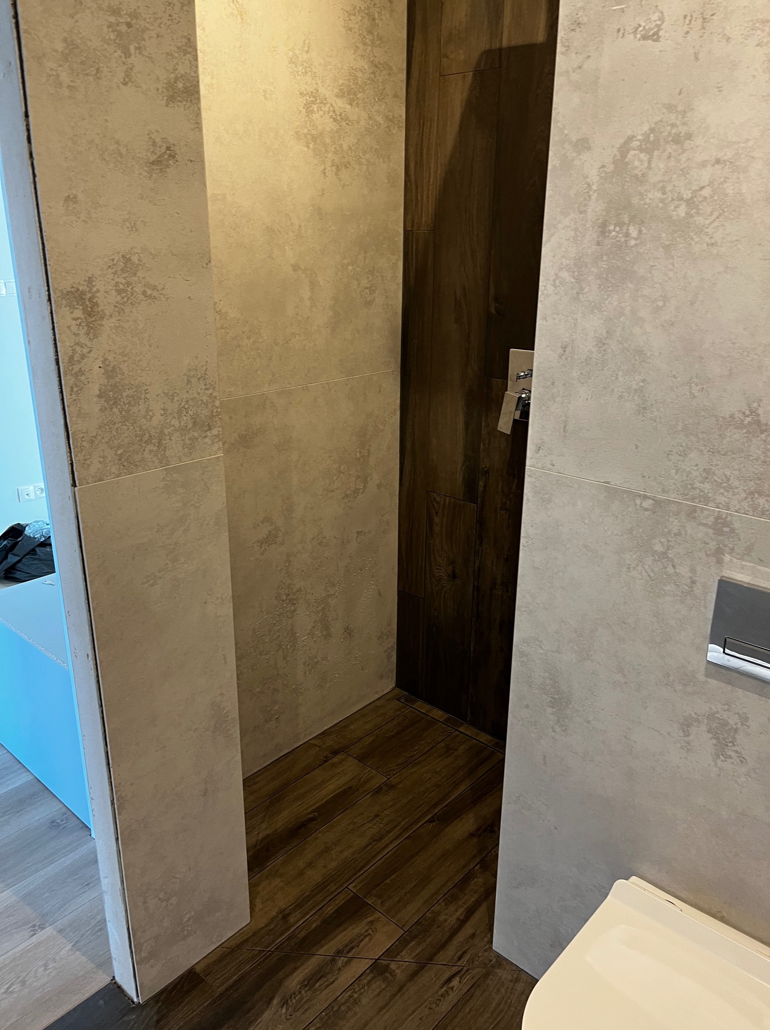 Wykończona kabina prysznicowa z drewnopodobną podłogą i szarą, teksturowaną okładziną ścienną. Widoczna bateria podtynkowa i fragment toalety.