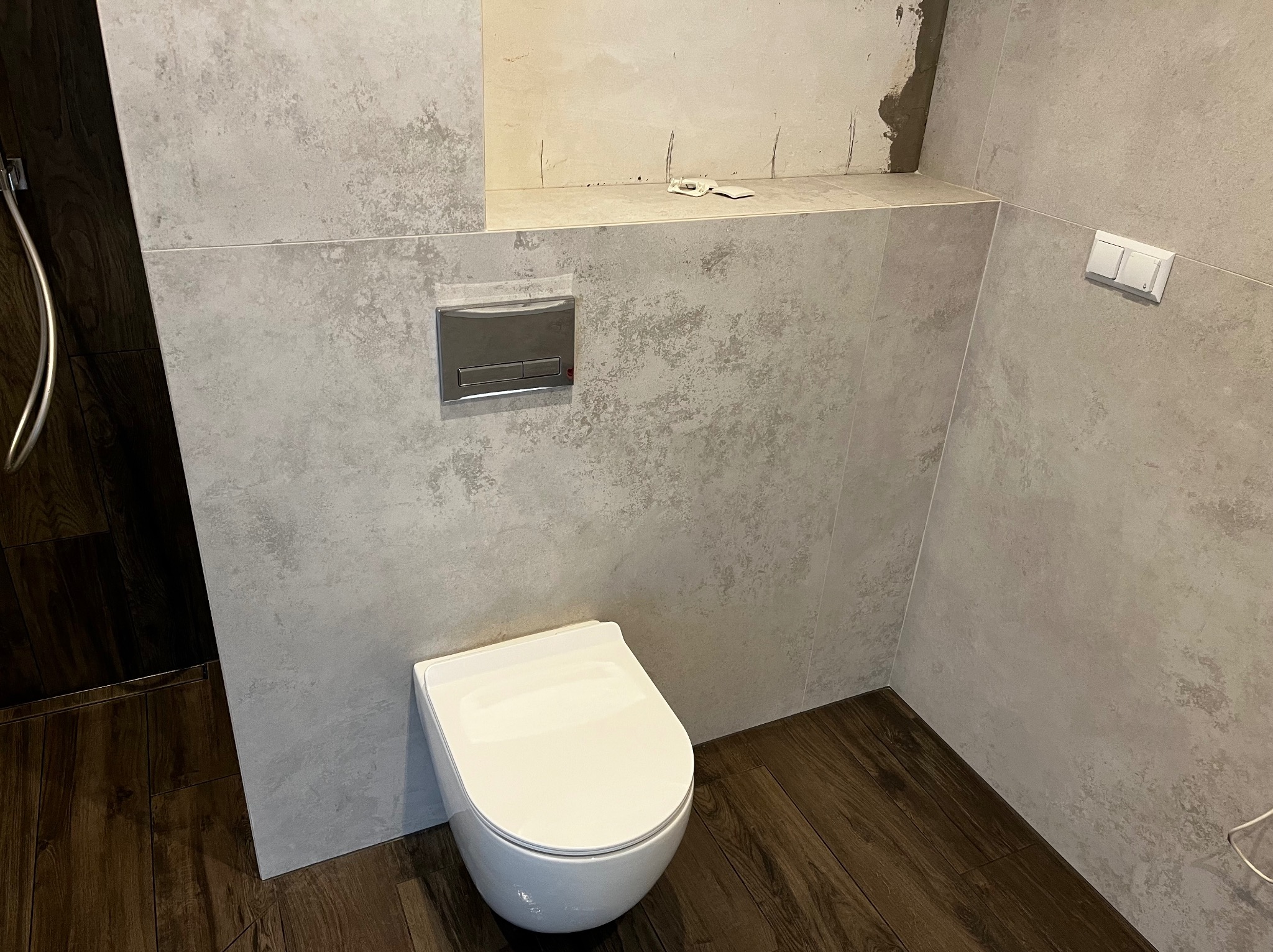 Nowoczesna, szara łazienka z podwieszaną toaletą i drewnopodobną podłogą. Widoczne płytki imitujące beton i chromowany przycisk spłuczki.