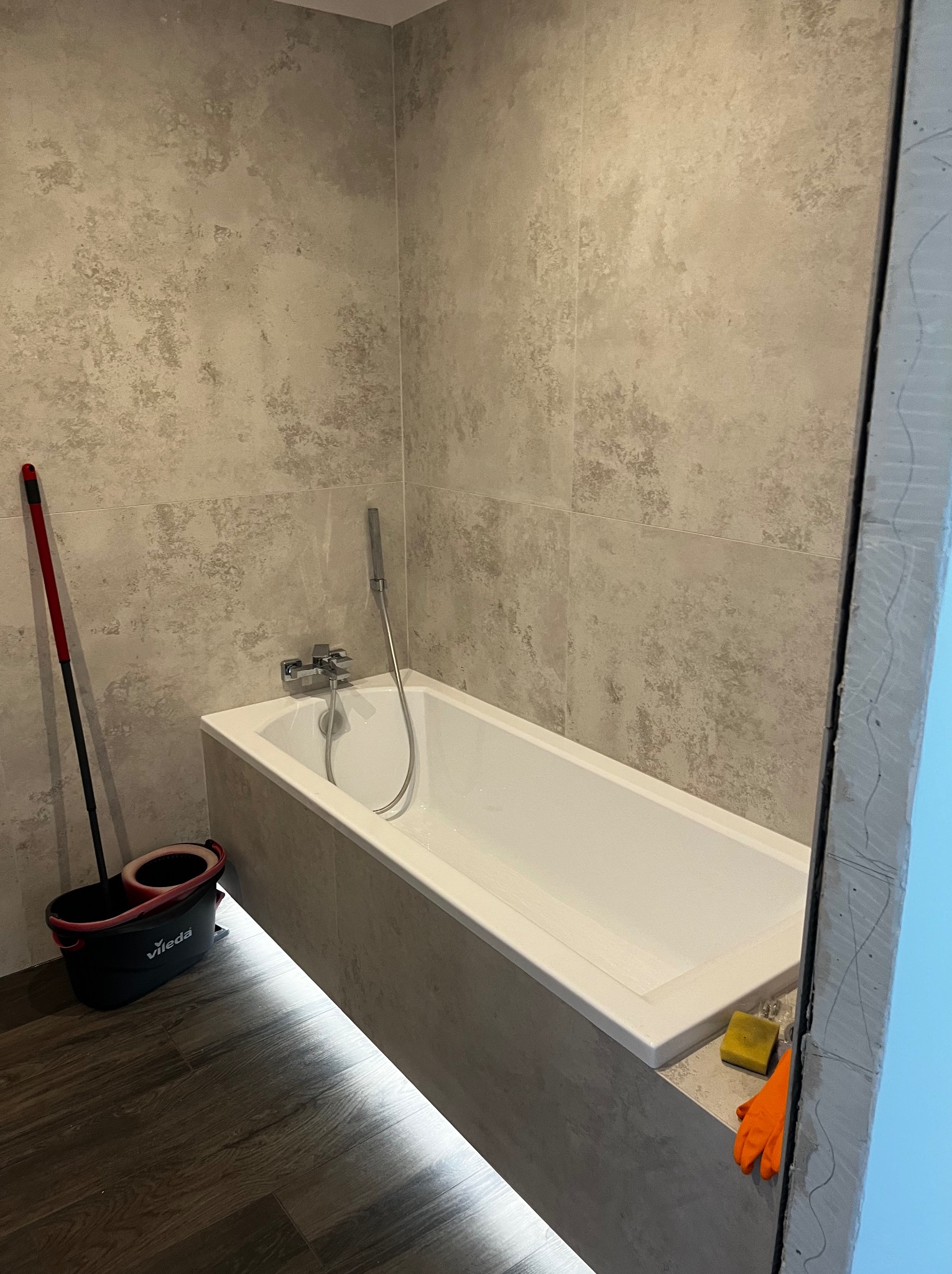 Wykończona łazienka w Konstancinie-Jeziornej: wanna obudowana płytkami, ściany wyłożone gresem imitującym kamień. Widoczne narzędzia pracy i wiadro z mopem.