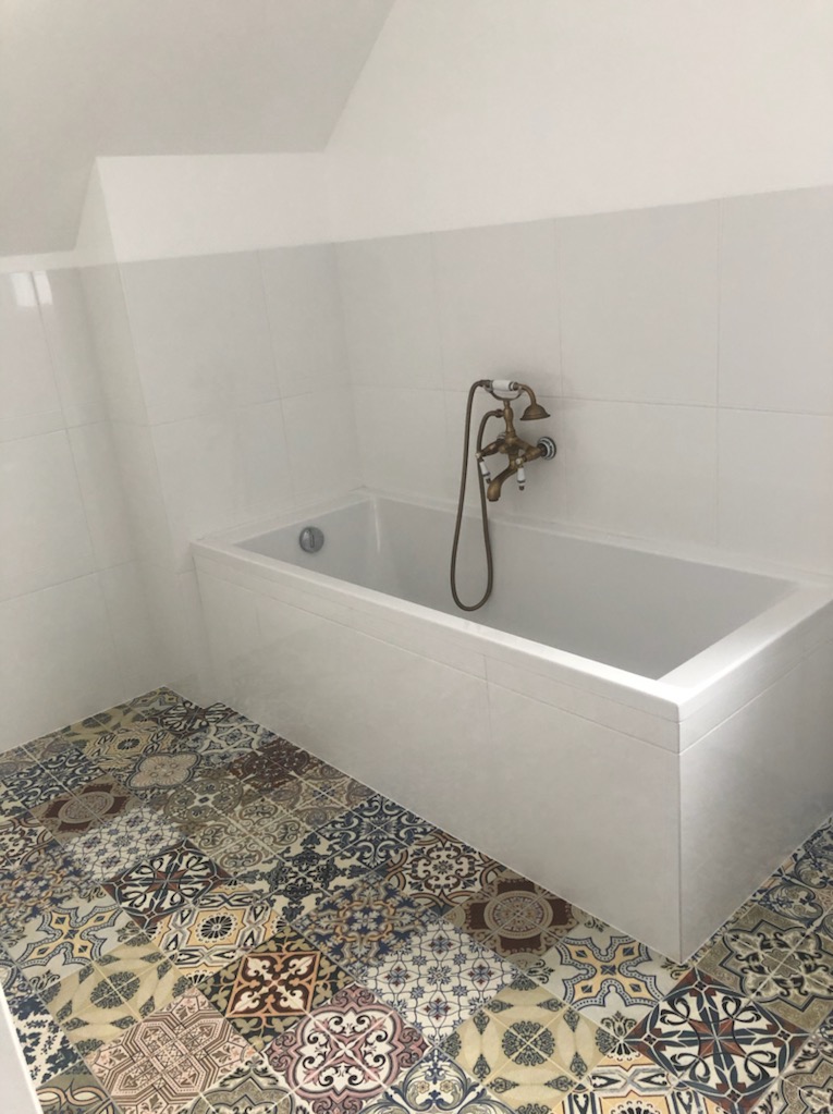 Biała wanna z baterią retro na tle białych płytek i wzorzystej podłogi w stylu patchwork, w jasnej łazience na poddaszu.