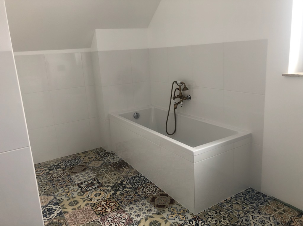 Biała wanna w łazience na poddaszu z patchworkową podłogą i armaturą w stylu retro. Ściany wyłożone białymi, kwadratowymi płytkami.