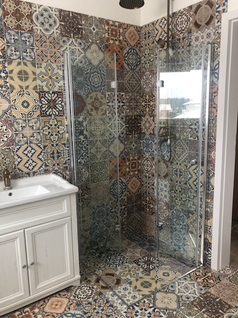 Wykończona łazienka z kabiną prysznicową za szkłem i umywalką w białej szafce. Ściany i podłoga wyłożone płytkami patchwork w ciepłych odcieniach. Czarna deszczownica.