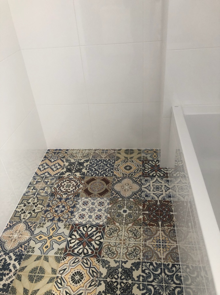 Wnętrze łazienki z dekoracyjną, wzorzystą podłogą z płytek ceramicznych. Białe, gładkie płytki na ścianach i fragment kabiny prysznicowej.