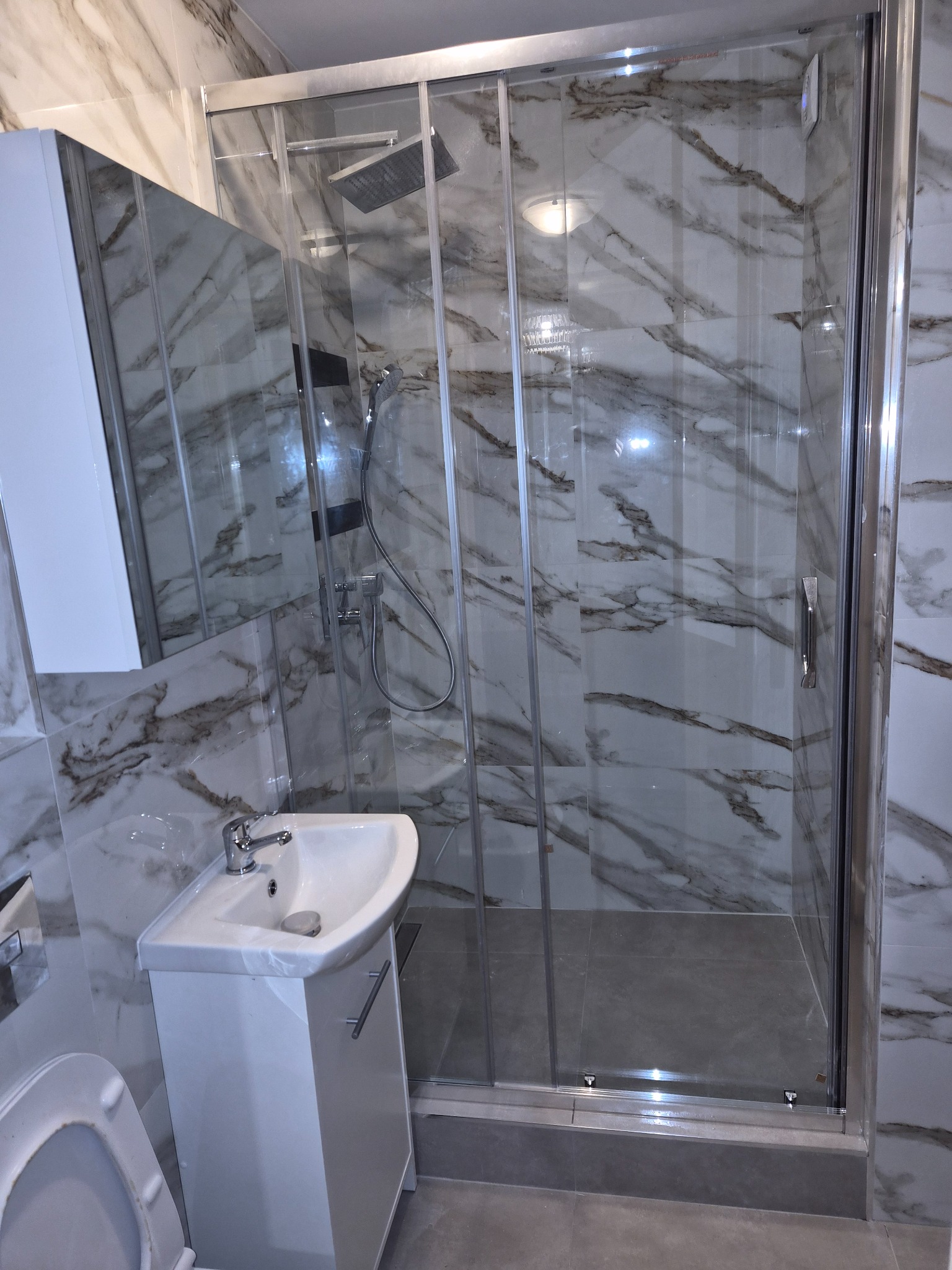 Wykończona łazienka z kabiną prysznicową ze szklanymi drzwiami przesuwnymi, umywalką z szafką i toaletą. Marmurowe płytki na ścianach i szara podłoga.