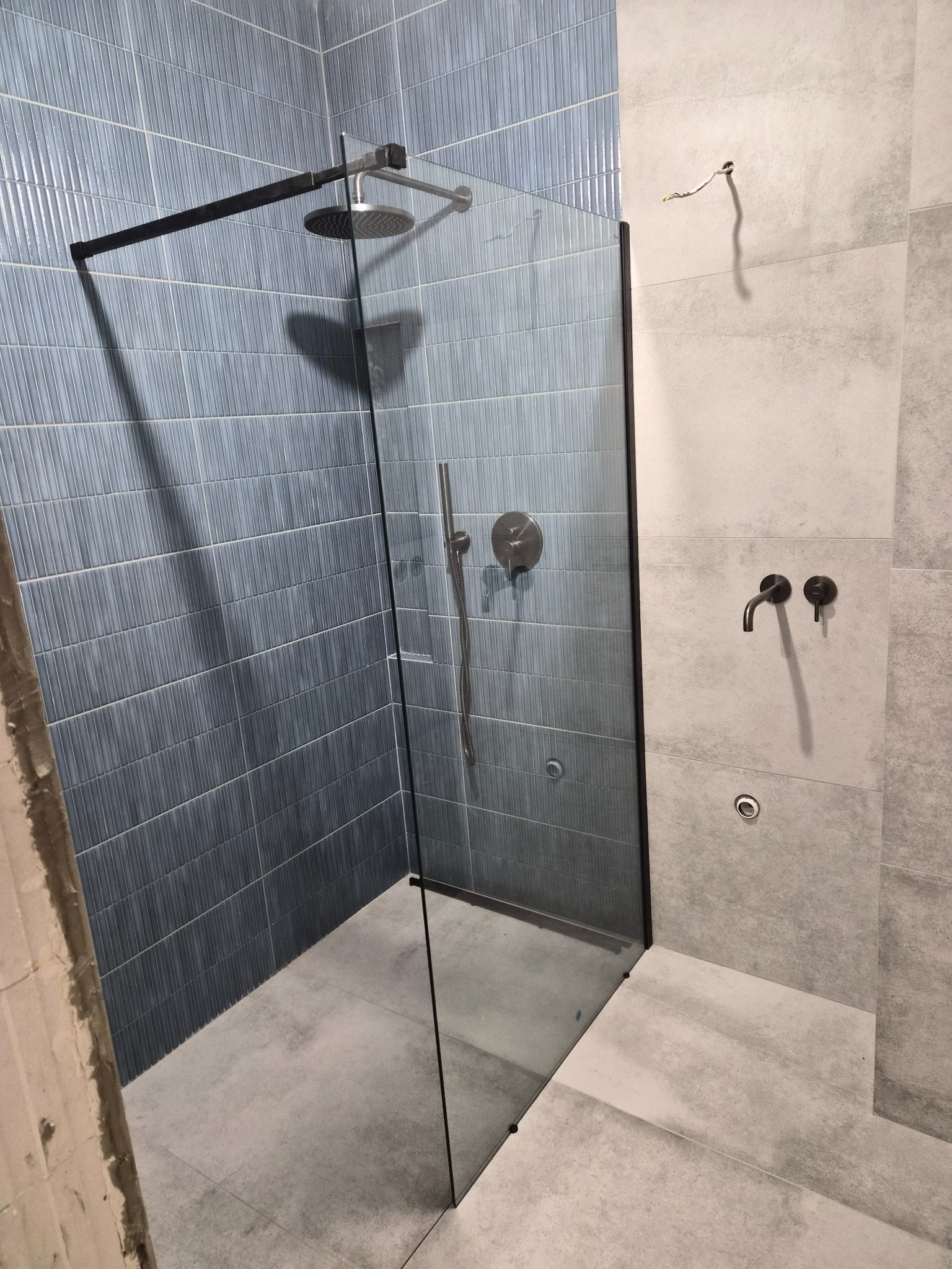 Kabina prysznicowa walk-in z czarną ramą i deszczownicą, wykończona niebieską mozaiką i szarą płytką. Widoczne baterie w kolorze czarnym. Minimalistyczny design.