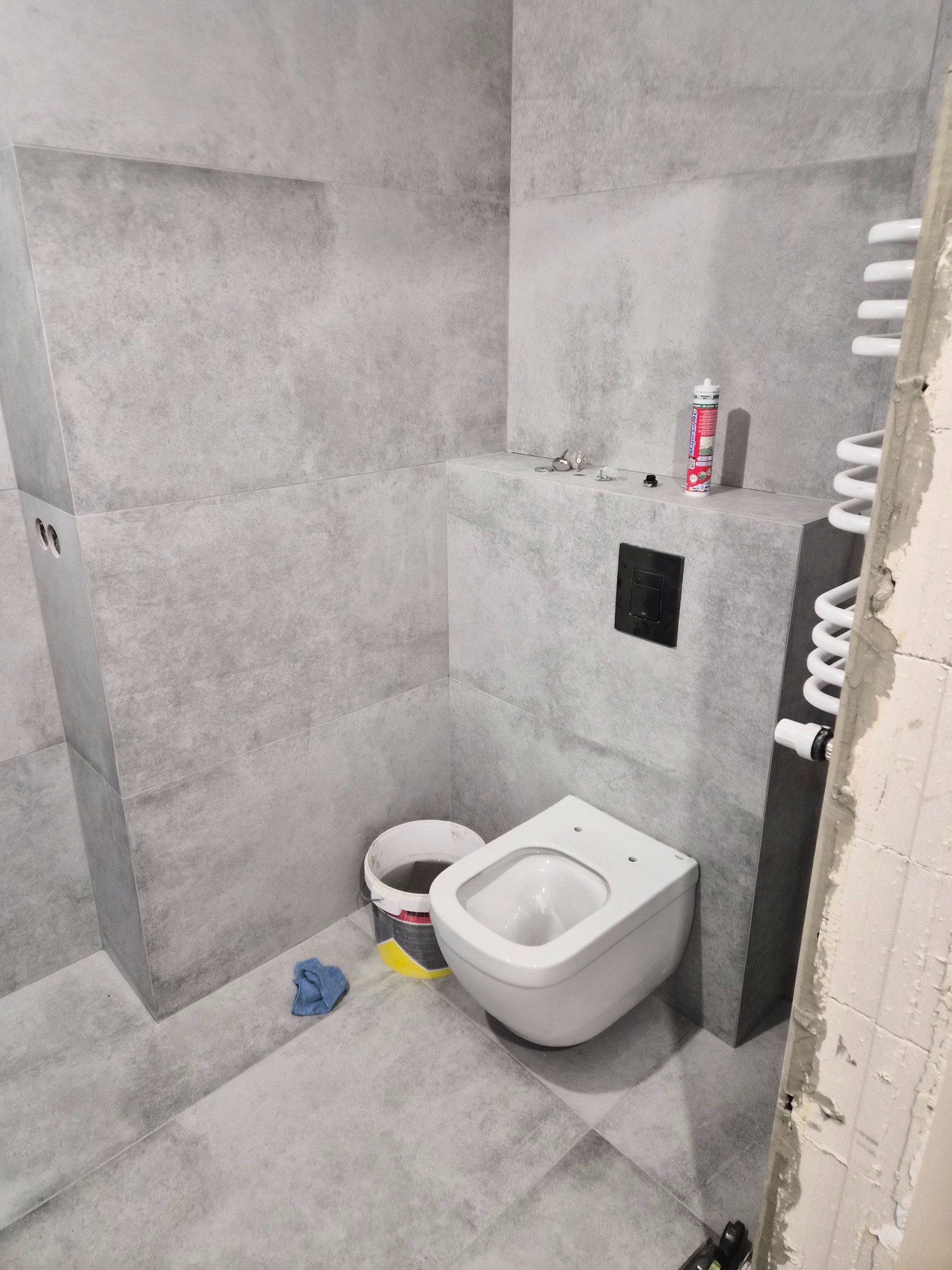 Szara łazienka w trakcie wykańczania: podwieszana toaleta, grzejnik drabinkowy, płytki imitujące beton, wiadro i akcesoria montażowe. Widoczne ślady prac wykończeniowych.