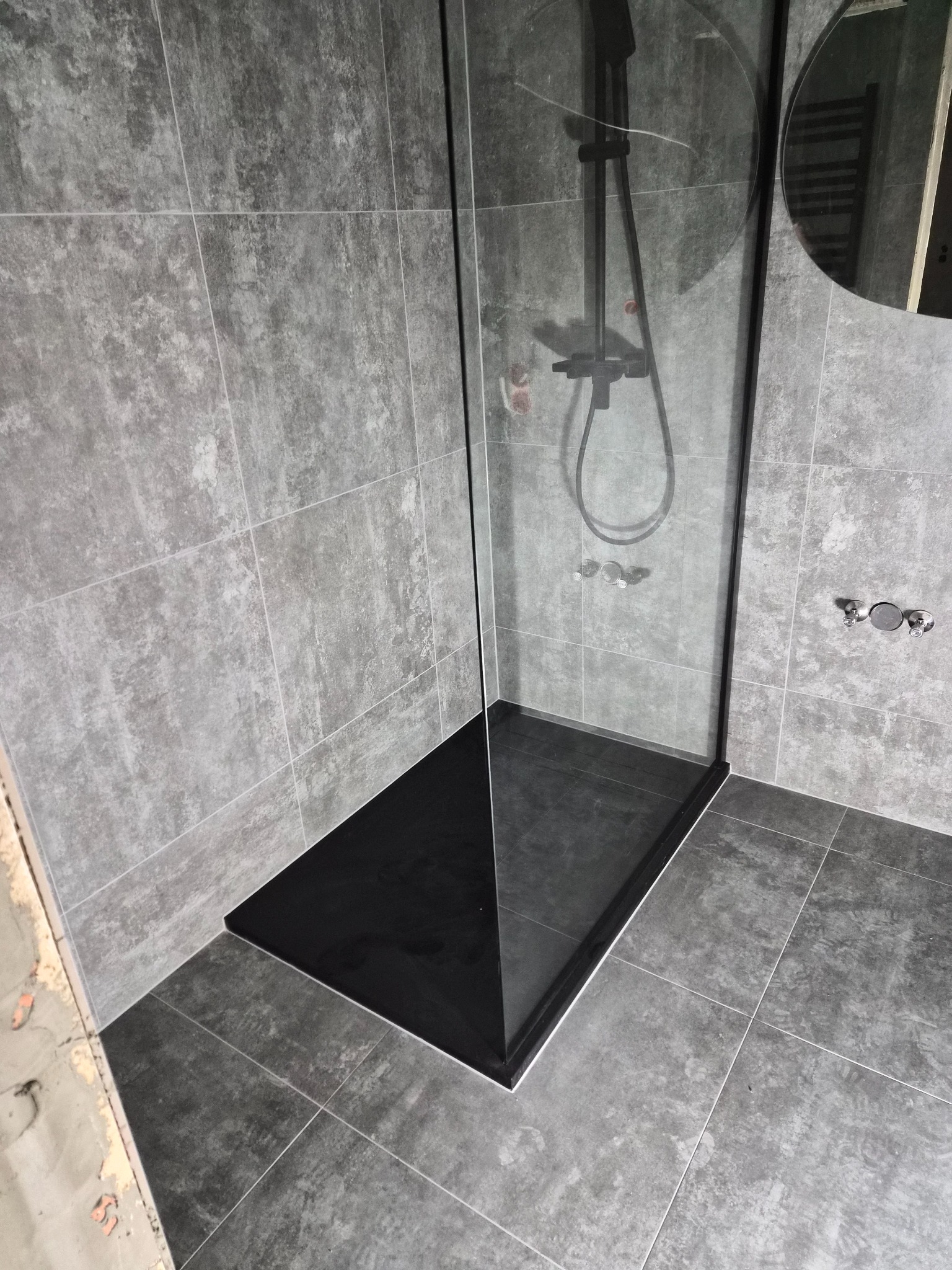 Nowoczesna kabina prysznicowa z czarnym brodzikiem i szklaną ścianką, szare płytki na ścianach i podłodze, minimalistyczny design, widoczne elementy instalacji.