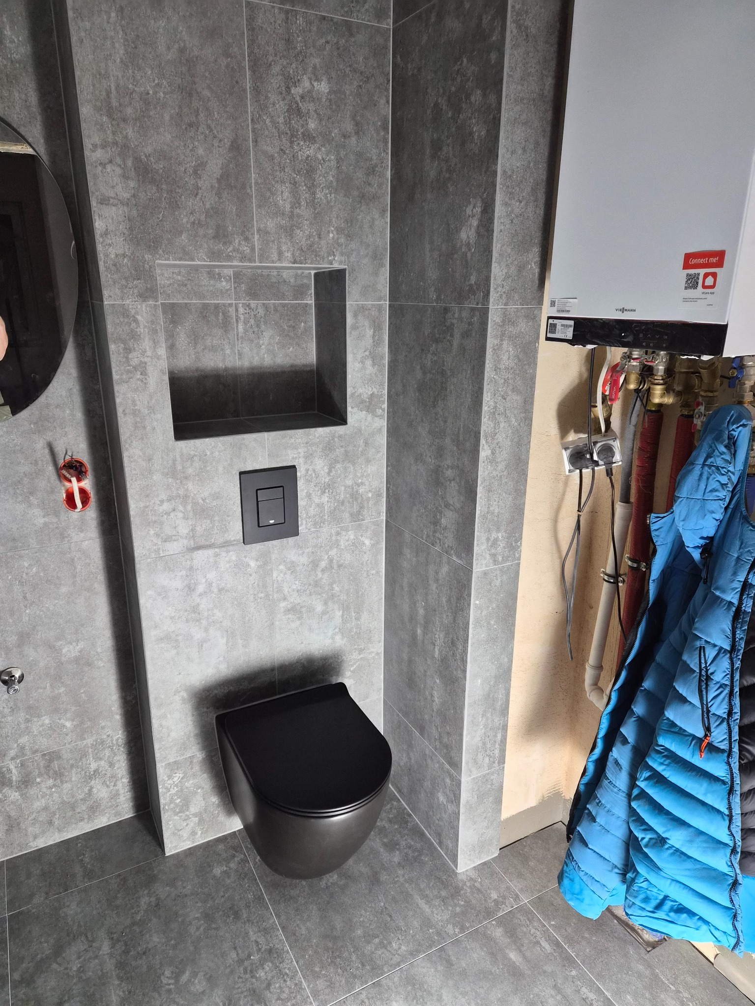 Nowoczesna łazienka z szarymi płytkami, czarną miską WC i ukrytą spłuczką. Na ścianie wnęka i wiszący piec gazowy. Błękitna kurtka dodaje koloru.