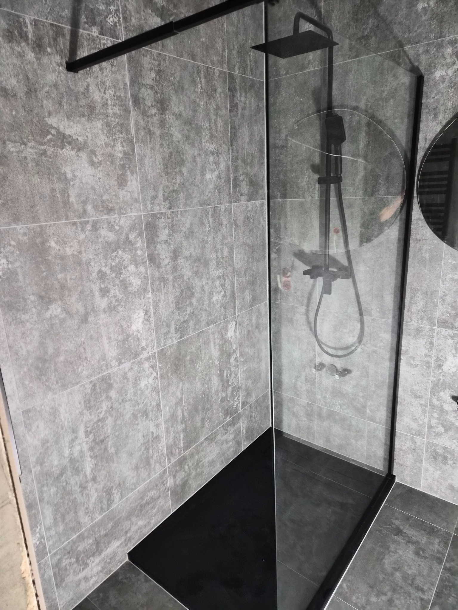 Nowoczesna kabina prysznicowa walk-in z czarną armaturą i szklaną ścianką na tle szarych, kwadratowych płytek. Minimalistyczny design, widoczne odbicie w szkle.