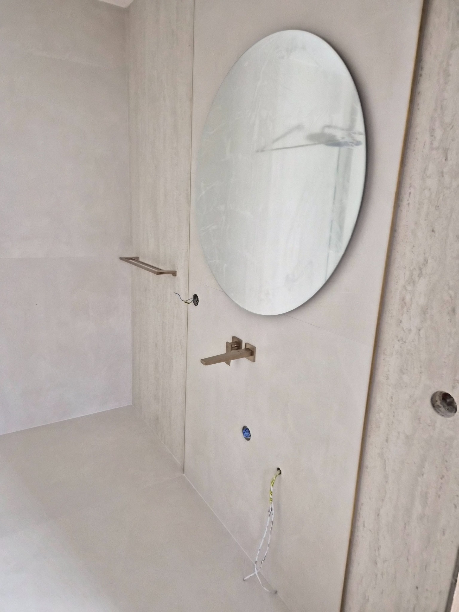 Minimalistyczna łazienka w odcieniach szarości z okrągłym lustrem, baterią i wieszakiem na ręczniki. Widoczne niedokończone podłączenia elektryczne.