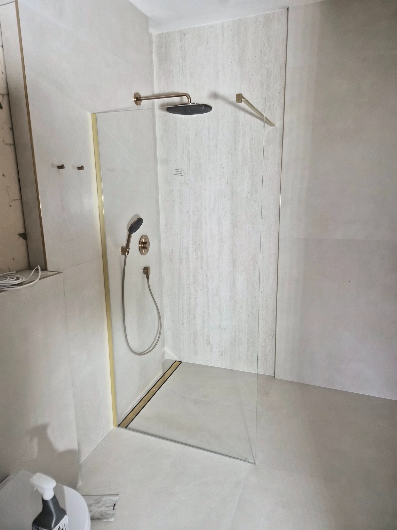 Wykończona kabina prysznicowa walk-in z elementami w kolorze złotym, zamontowana w łazience wykończonej w jasnych płytkach. Widoczny odpływ liniowy.