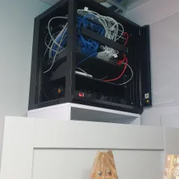 Otwarta szafa rack z okablowaniem sieciowym, umieszczona na białej szafce, z drewnianą drabiną malarską opartą o ścianę z prawej strony.