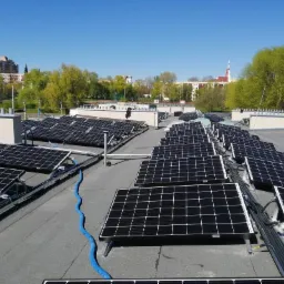 Rozległa instalacja fotowoltaiczna na płaskim dachu budynku, widoczne moduły, kable i niebieski przewód, w tle zabudowa miejska i błękitne niebo.