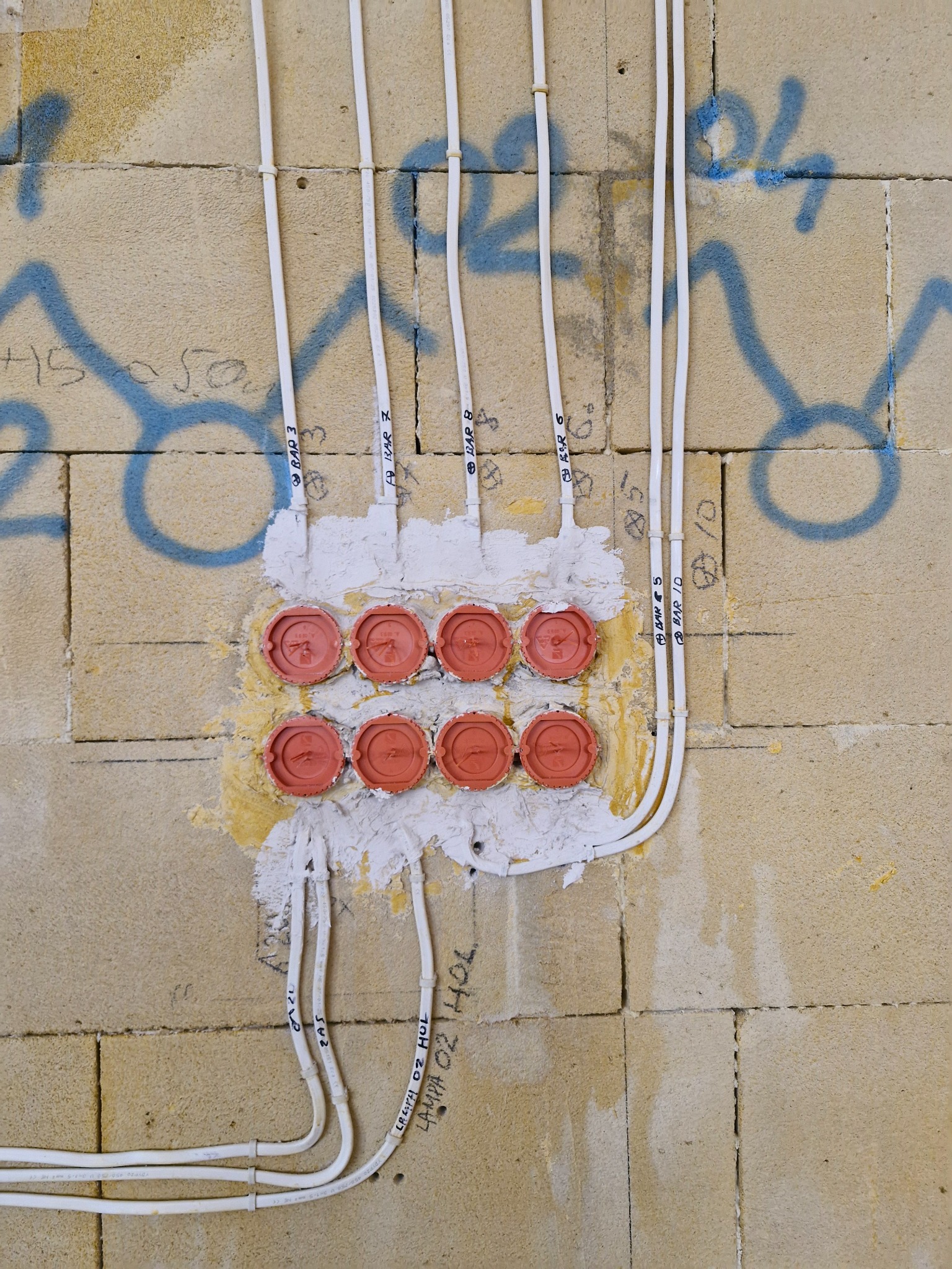 Niedokończona instalacja elektryczna: białe kable prowadzone po surowej ścianie z betonu komórkowego z widocznymi napisami i niebieskimi graffiti, osiem czerwonych puszek instalacyjnych...