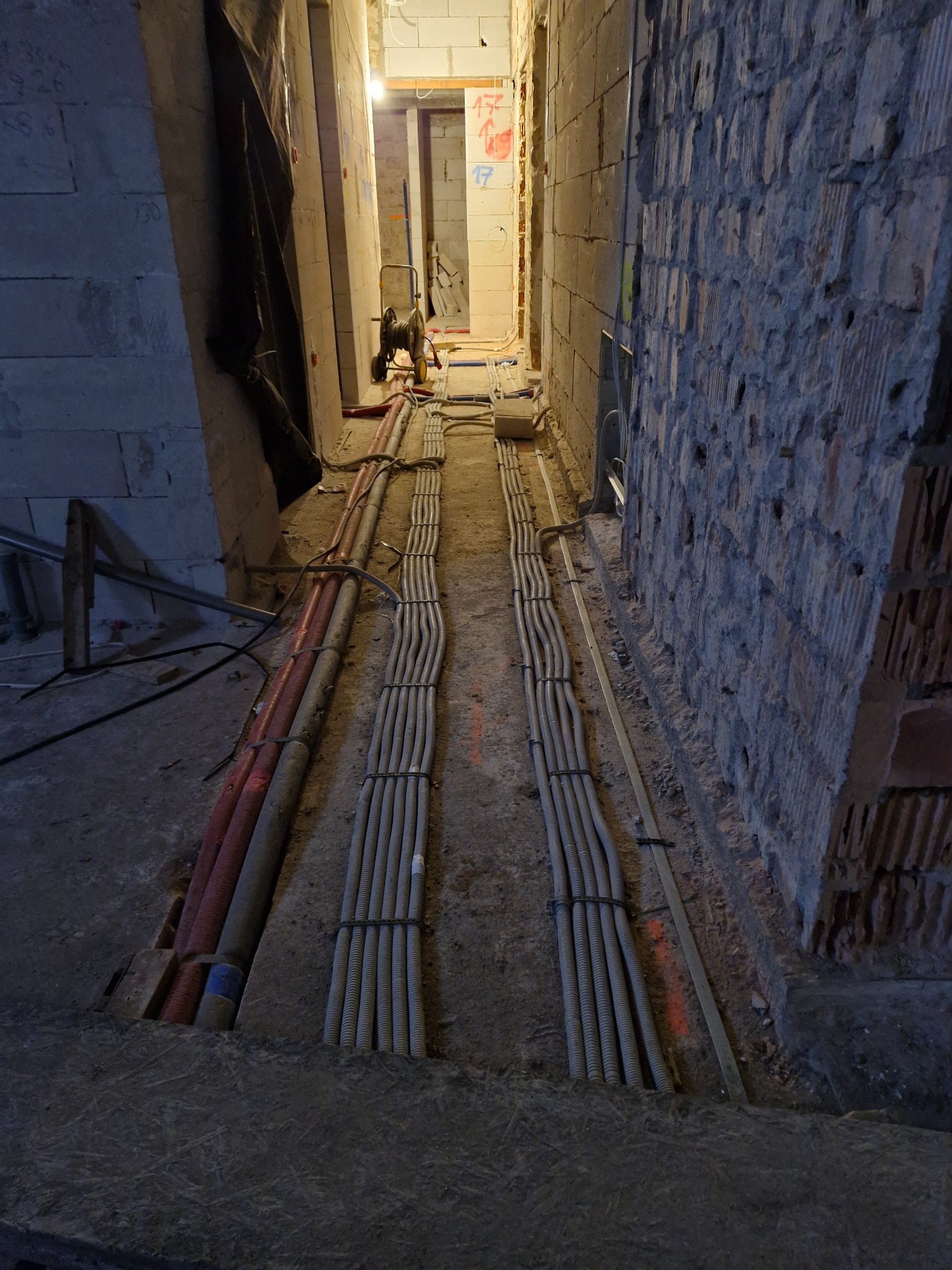 Wąski korytarz w surowym stanie z widocznymi przewodami elektrycznymi ułożonymi w korytkach podłogowych, ściany z cegły i bloczków, instalacja w trakcie montażu.