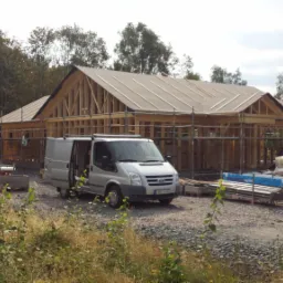Wznoszenie konstrukcji domu z drewna, widoczny szkielet budynku z deskowaniem dachu, na pierwszym planie samochód dostawczy i materiały budowlane.