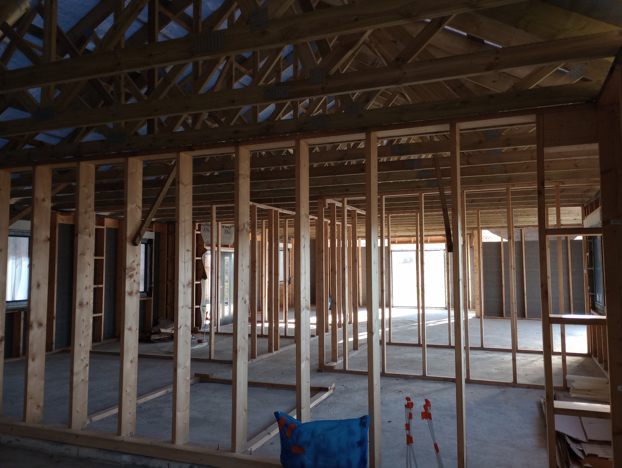 Wnętrze domu w trakcie budowy w technologii szkieletowej, widoczne drewniane ramy ścian i stropu, podłoga betonowa, niebieska poduszka i statyw na podłodze.