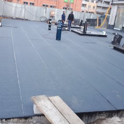 REGION BUD SP. ZO. O. - Montaż izolacji na dachu płaskim z widocznymi pracownikami, butlą gazową i narzędziami budowlanymi w tle, w trakcie realizacji.