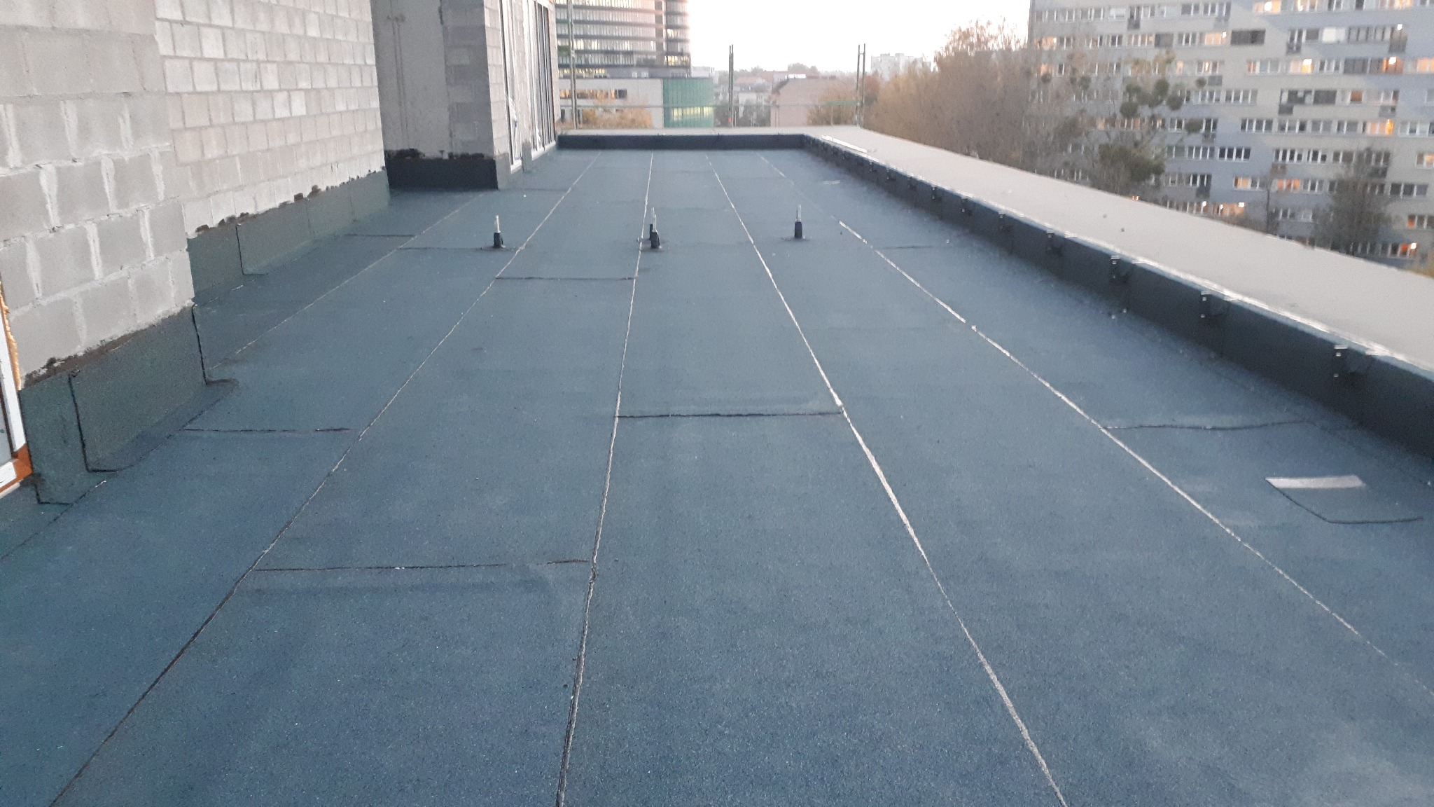 Płaski dach pokryty papą termozgrzewalną z widocznymi odpowietrznikami, budynki mieszkalne w tle.