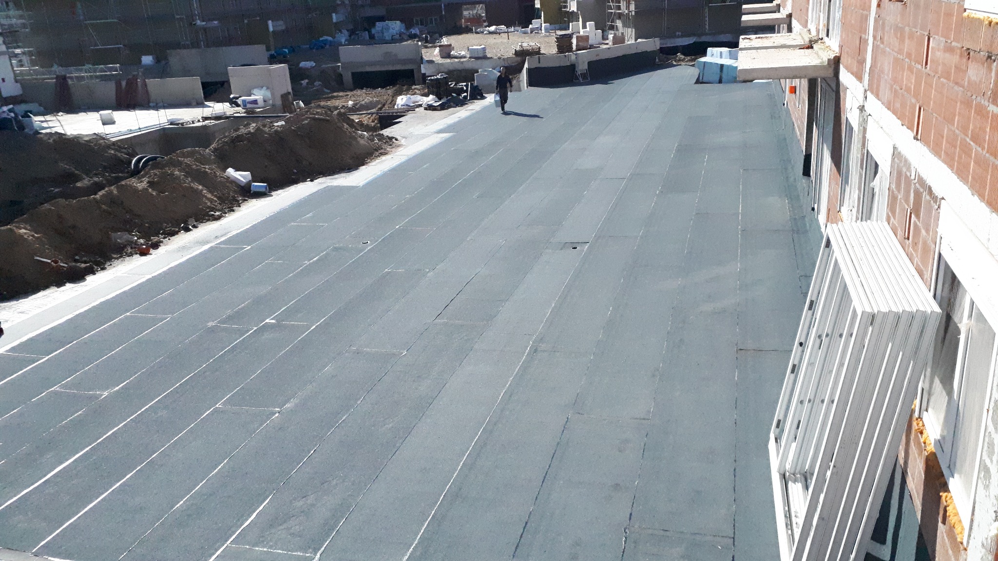Nowe krycie dachu papą termozgrzewalną na budynku w trakcie budowy, widoczna osoba w oddali i okna czekające na montaż.