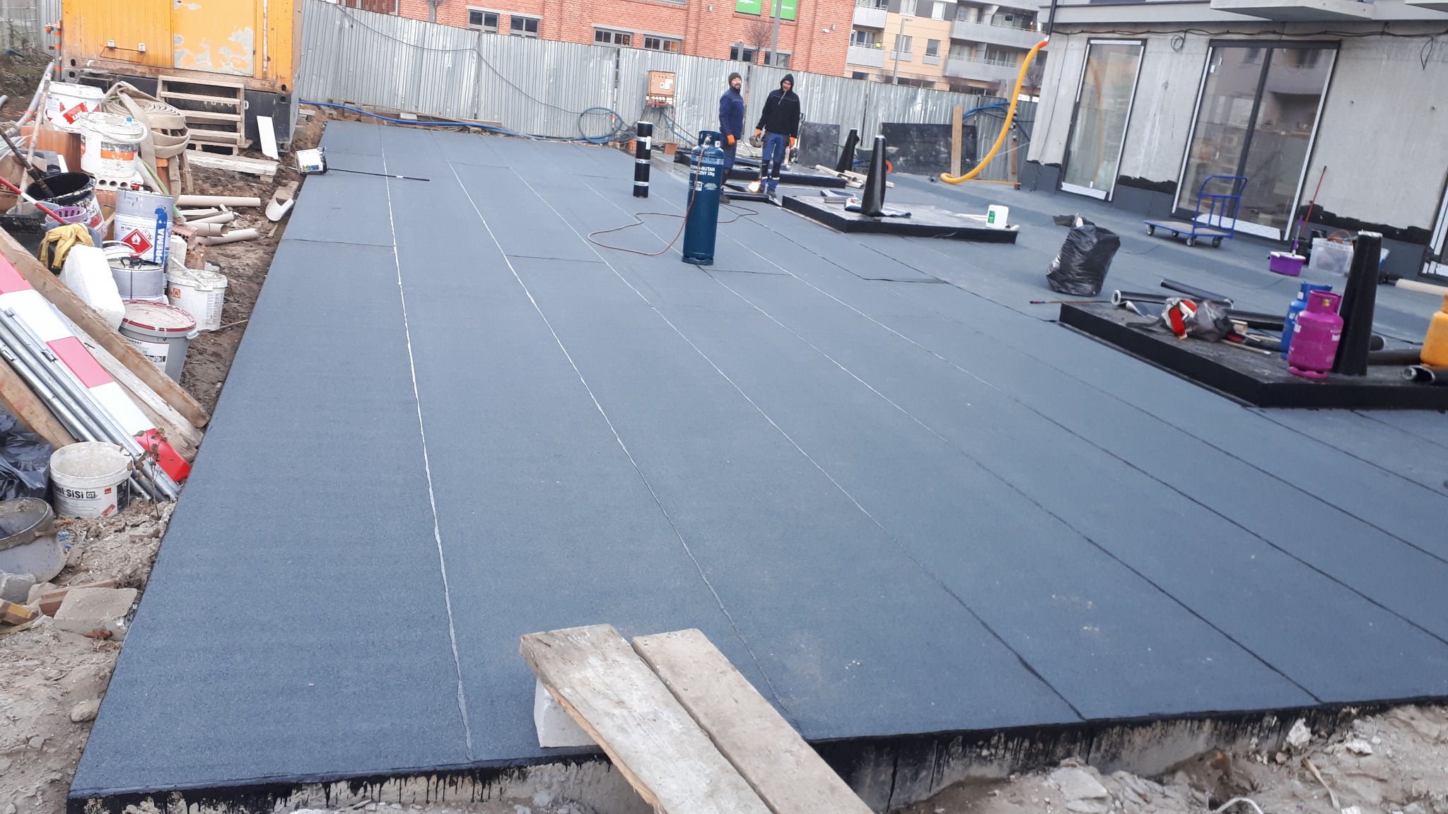 Montaż izolacji na dachu płaskim z widocznymi pracownikami, butlą gazową i narzędziami budowlanymi w tle, w trakcie realizacji.