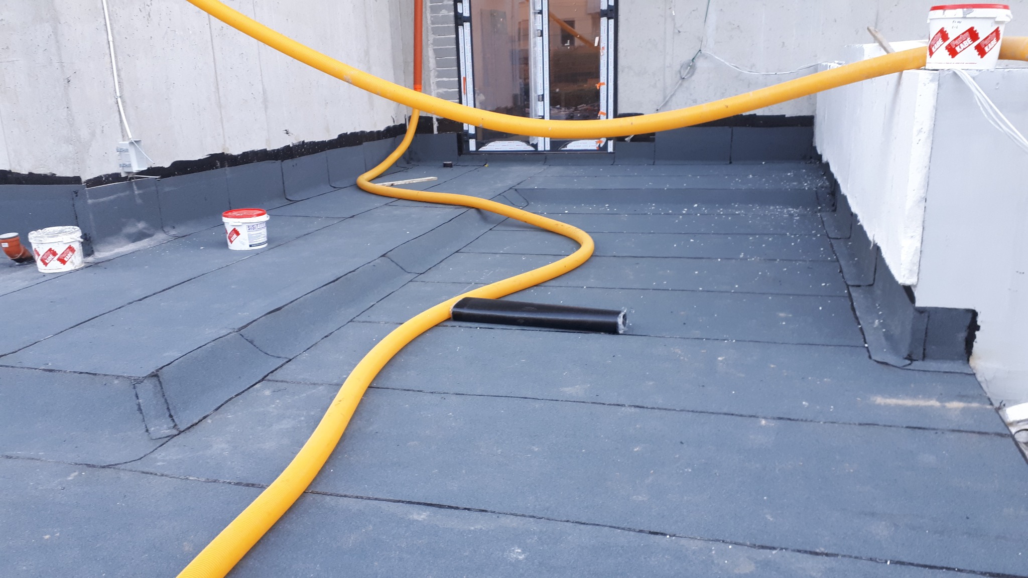 Nowo położona, płaska powierzchnia dachu pokryta papą, z widoczną żółtą rurą wentylacyjną, rolką papy i pojemnikami z masą bitumiczną w tle.