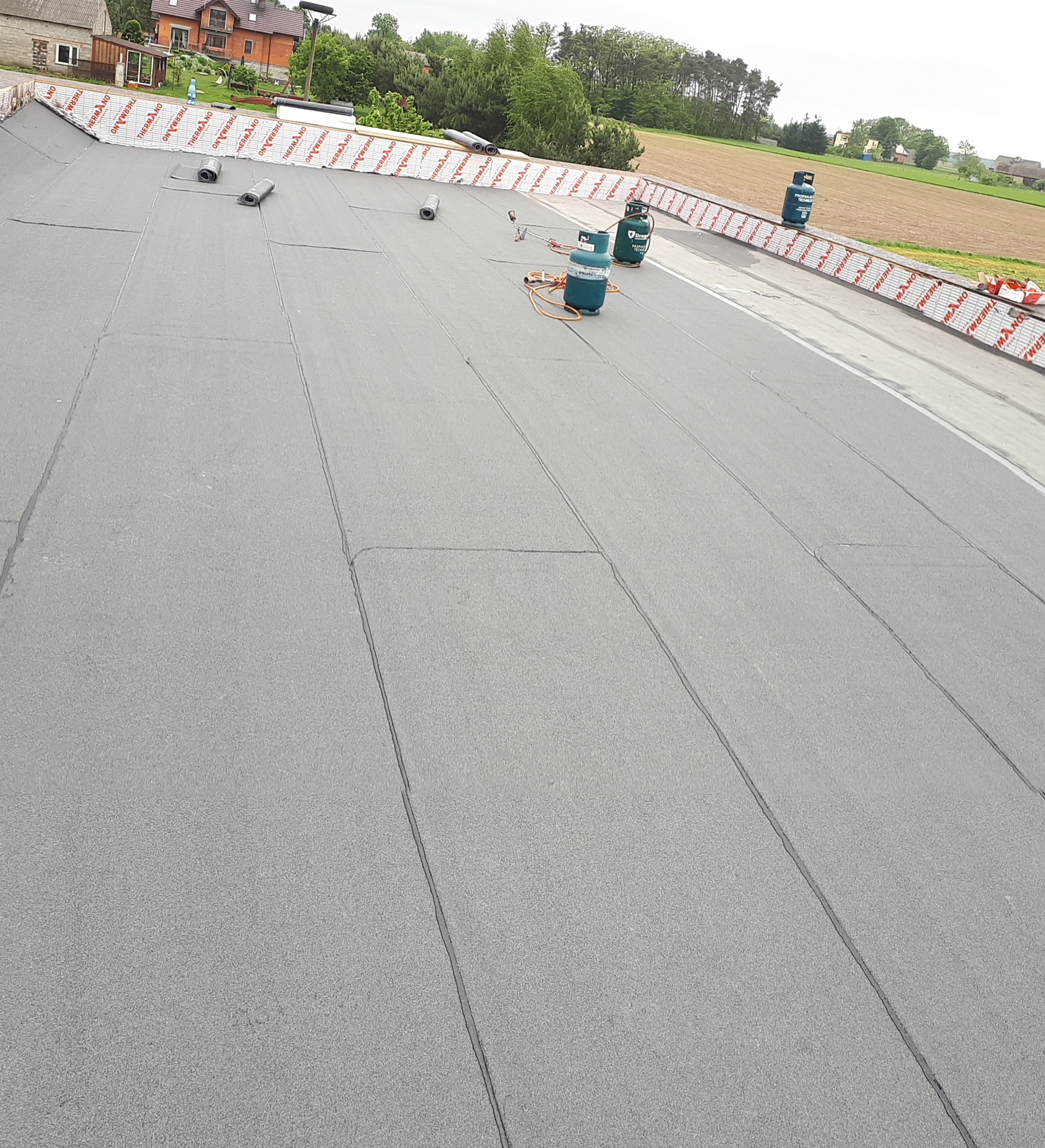 Widok z góry na płaski dach pokryty szarą papą termozgrzewalną, z widocznymi zgrzewami i rolkami papy wzdłuż krawędzi, butle gazowe i palnik dekarski na powierzchni, w tle budynki i pola.