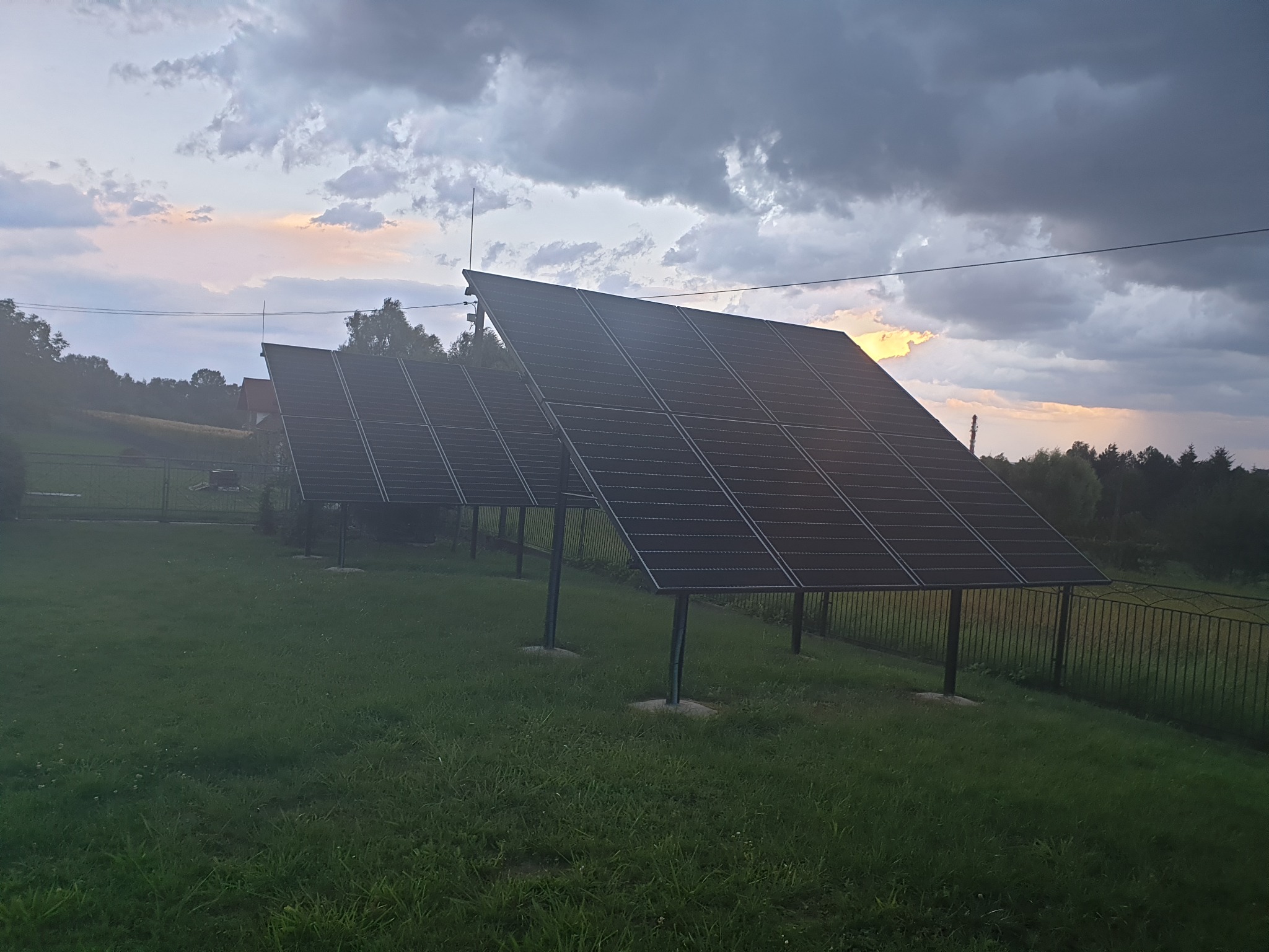 Dwa rzędy paneli słonecznych ustawione na trawiastym terenie, w tle zachodzące słońce przebijające przez ciemne chmury.