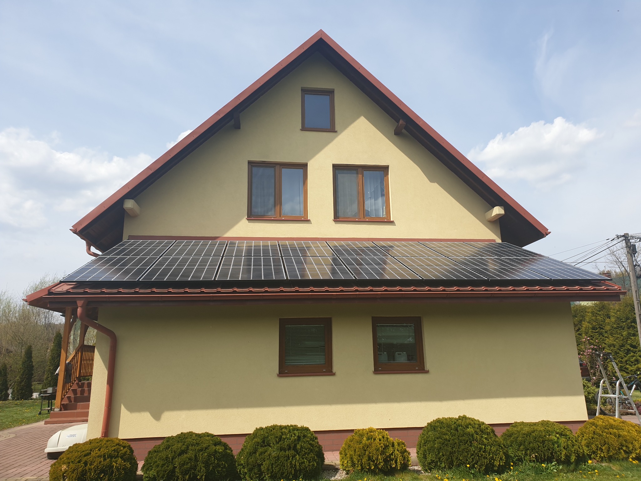 Dom jednorodzinny z panelami słonecznymi zamontowanymi na dachu, widok z niskiej perspektywy, błękitne niebo z chmurami w tle.