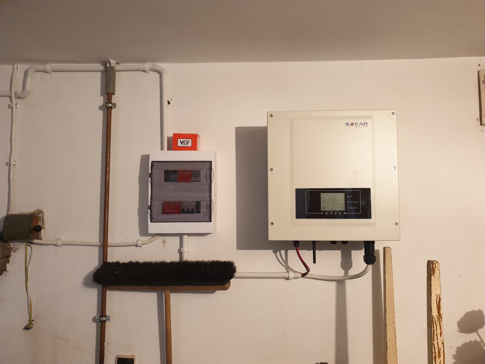 Biała skrzynka rozdzielcza i falownik SOFAR na ścianie, widoczne przewody, rury i stara miotła oparta o ścianę.