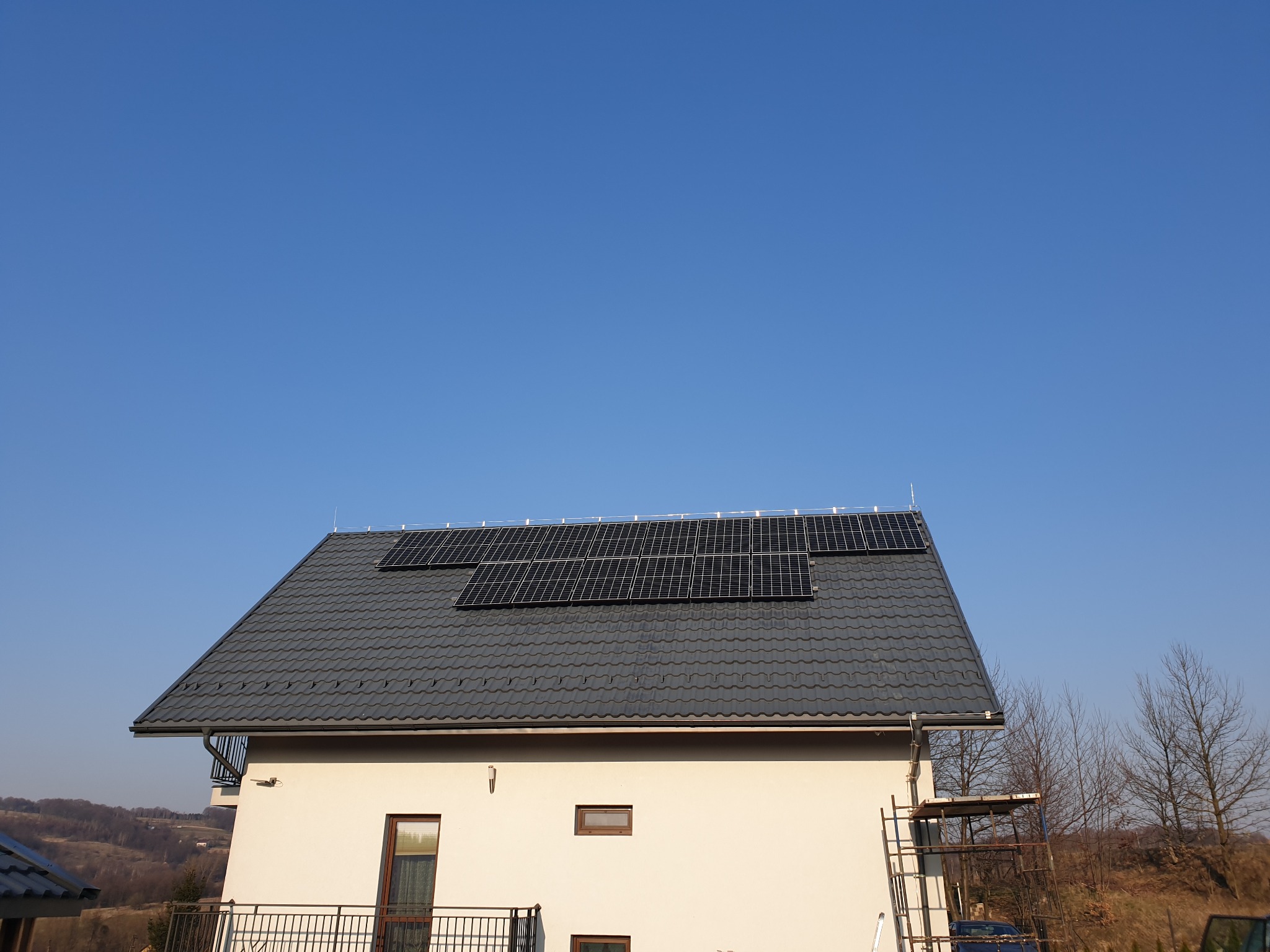 Panele słoneczne na dachu domu jednorodzinnego z szarą dachówką, widoczne zabezpieczenia odgromowe i drabina oparta o ścianę.