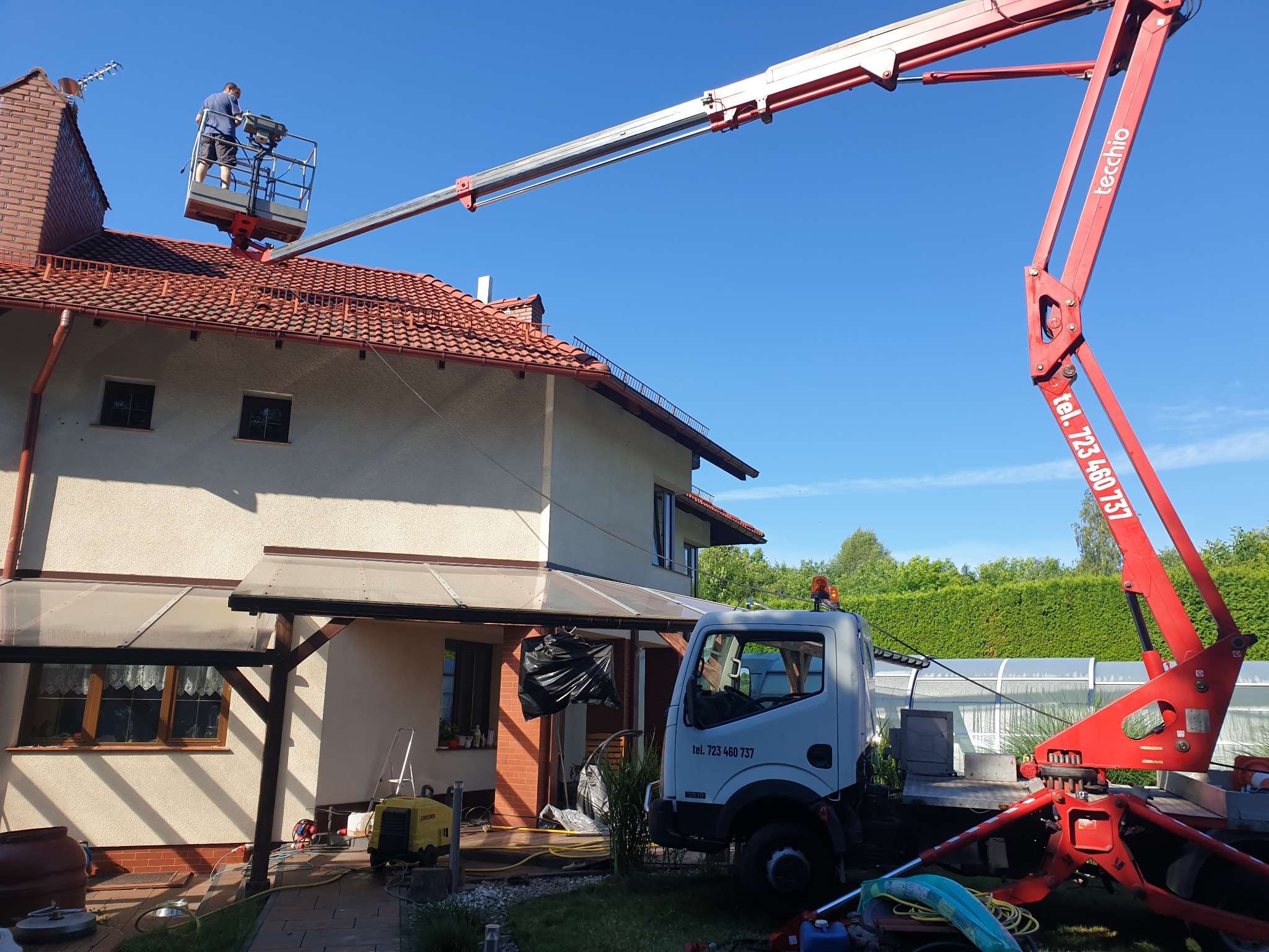 Pracownik na podnośniku koszowym przy dachu domu jednorodzinnego z ceglastą dachówką, w tle błękitne niebo i zielona roślinność.