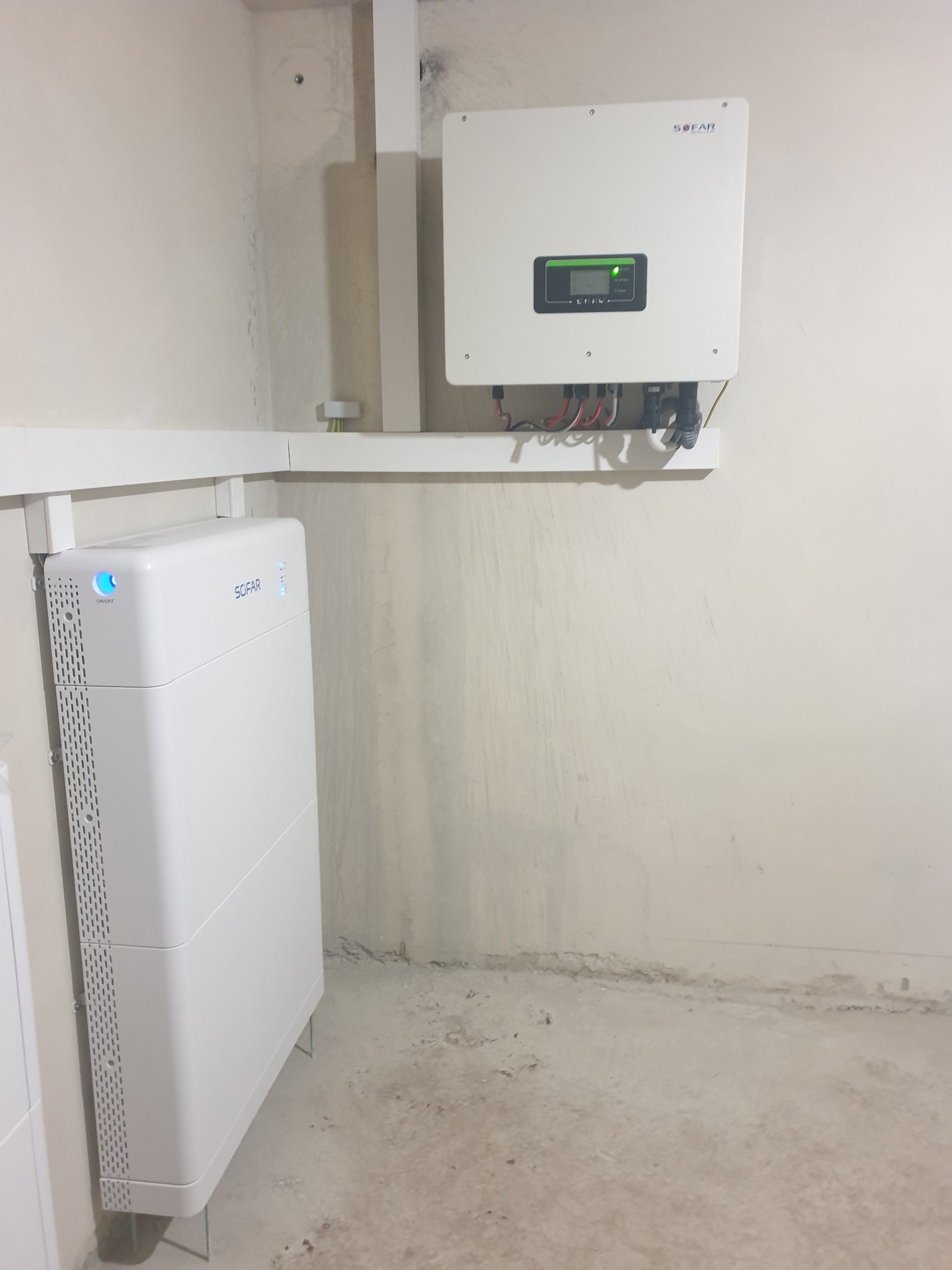 Biały inwerter i akumulator energii słonecznej marki SOFAR zamontowane na ścianie, widoczne kable i przewody, surowe wykończenie wnętrza.