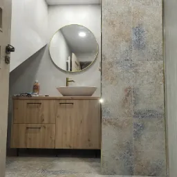 Nowoczesna łazienka z umywalką nablatową, okrągłym lustrem w złotej ramie, szafką z drewnopodobnym frontem i dekoracyjnymi płytkami na ścianie oraz podłodze.