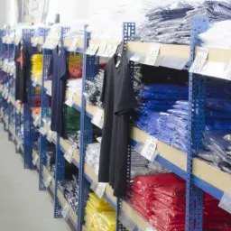 Hurtownia odzieży: rzędy niebieskich regałów wypełnione ułożonymi w stosy, zapakowanymi koszulkami w różnych kolorach, z zawieszonymi pojedynczymi egzemplarzami na wieszakach i etykietami cenowymi.