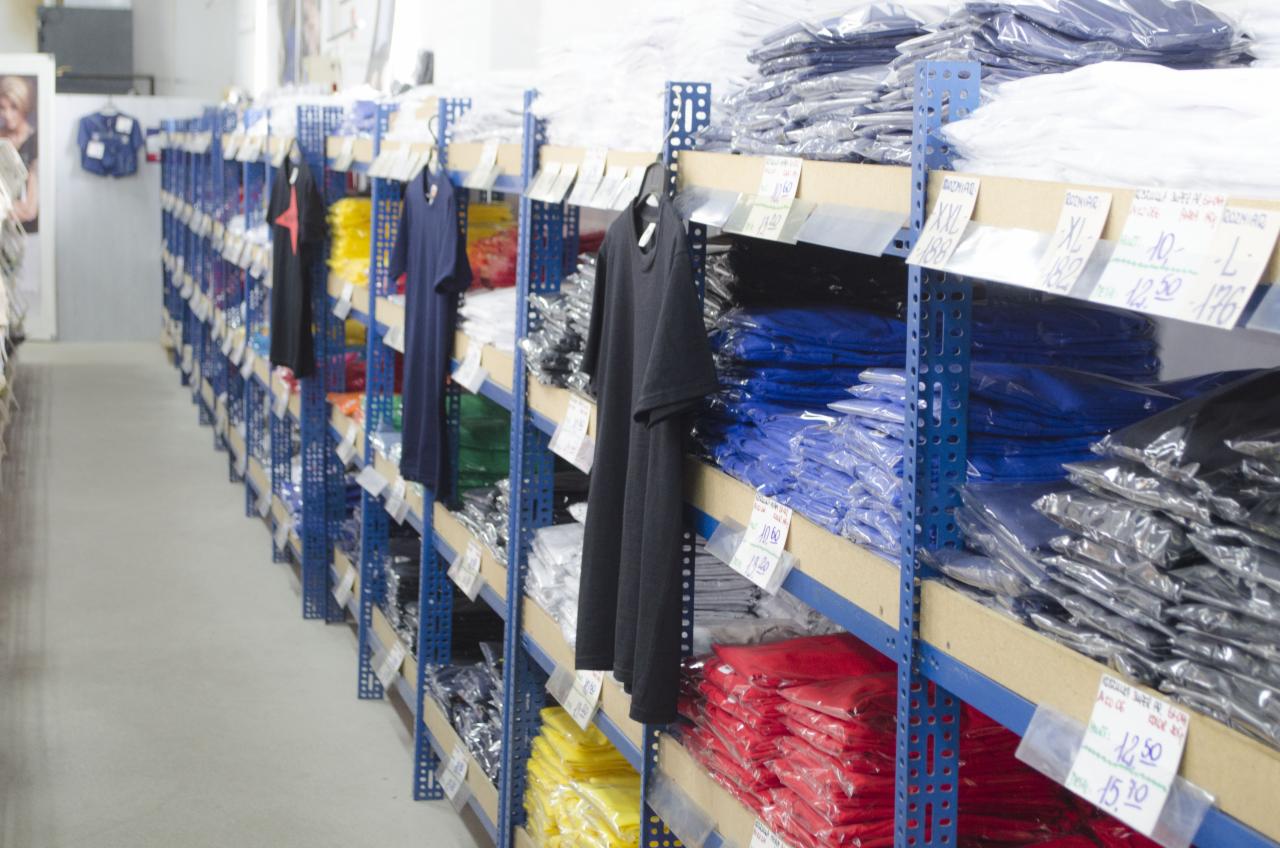 Hurtownia odzieży: rzędy niebieskich regałów wypełnione ułożonymi w stosy, zapakowanymi koszulkami w różnych kolorach, z zawieszonymi pojedynczymi egzemplarzami na wieszakach i etykietami cenowymi.