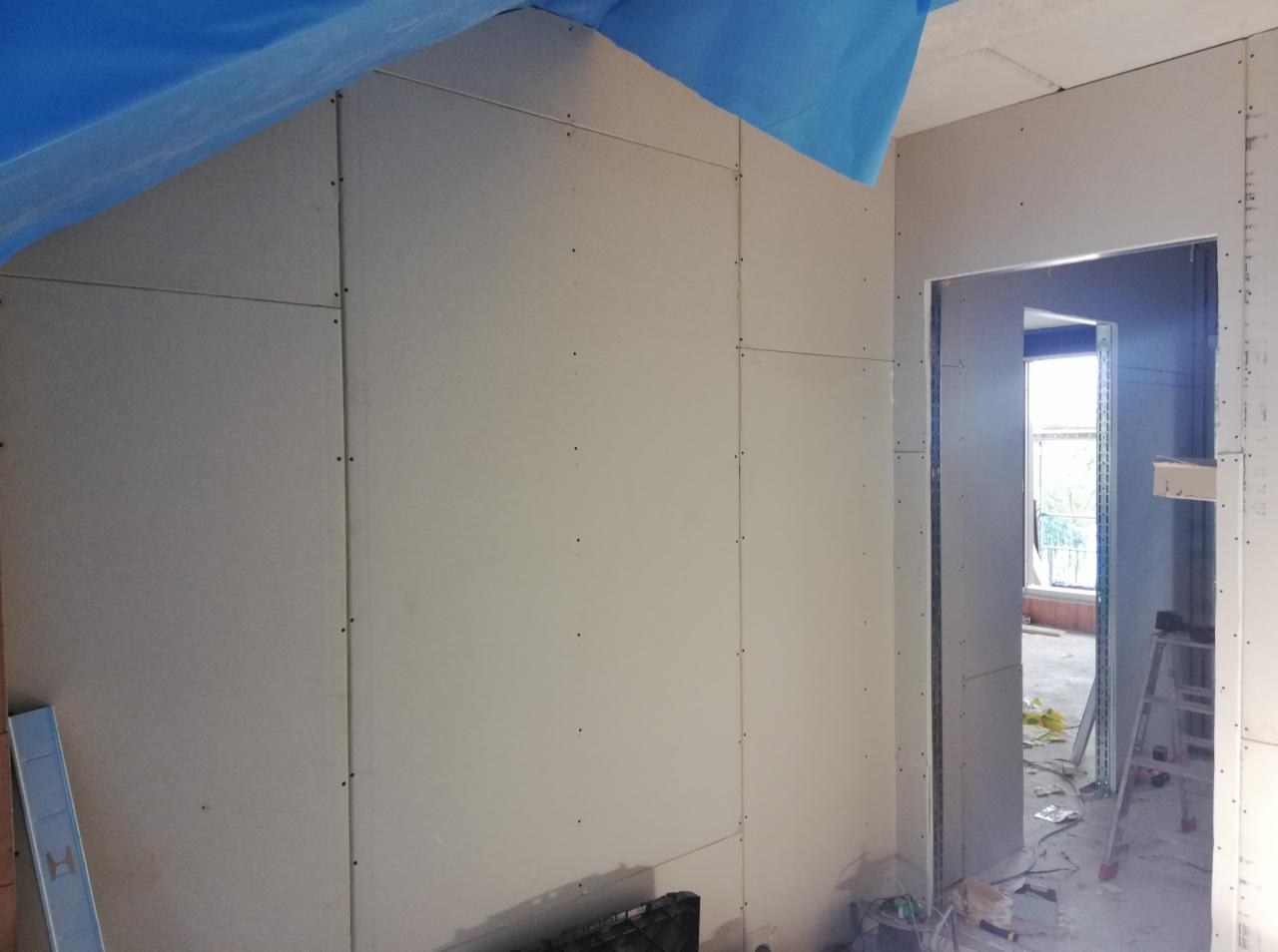 Montaż płyt kartonowo-gipsowych na ścianie i suficie pod skosem, widoczne łączenia skręcone wkrętami, zabezpieczenie folią malarską na suficie, poziomica w rogu pomieszczenia, widok na okno w oddali.