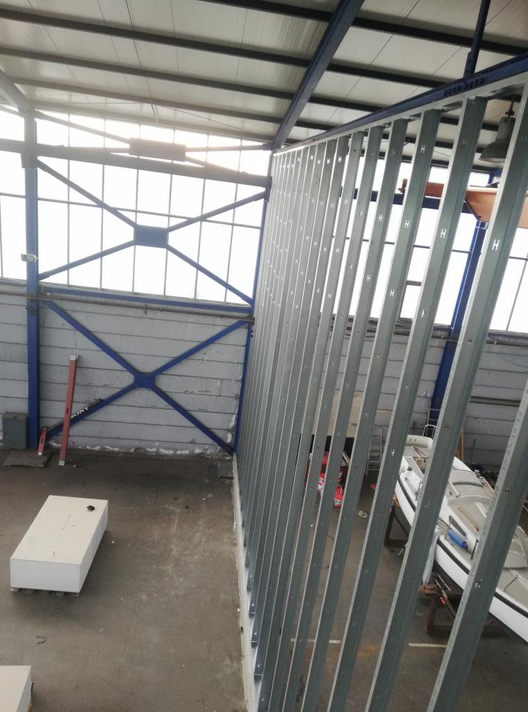 Stelaż z profili stalowych, prawdopodobnie pod ścianę działową, w hali przemysłowej z widocznymi elementami konstrukcyjnymi i oknami w tle.